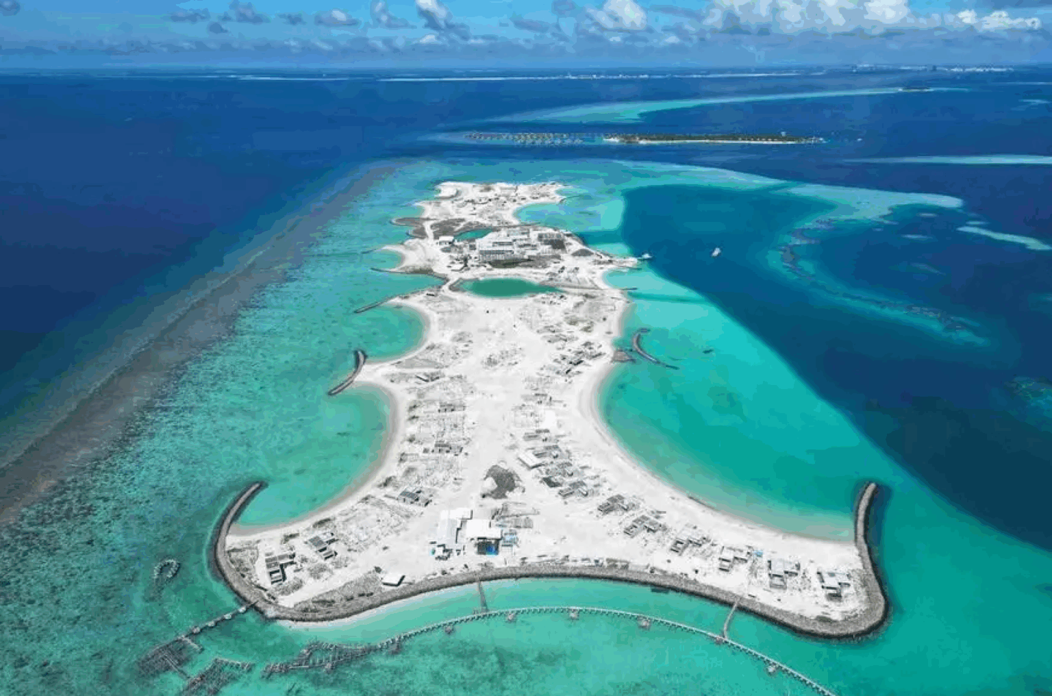 Aerial view of a bright, sandy island surrounded by turquoise and deep blue waters. Sparse buildings and construction are visible, with curved breakwaters extending into the sea. Fluffy clouds dot the clear blue sky, creating a serene, expansive atmosphere.