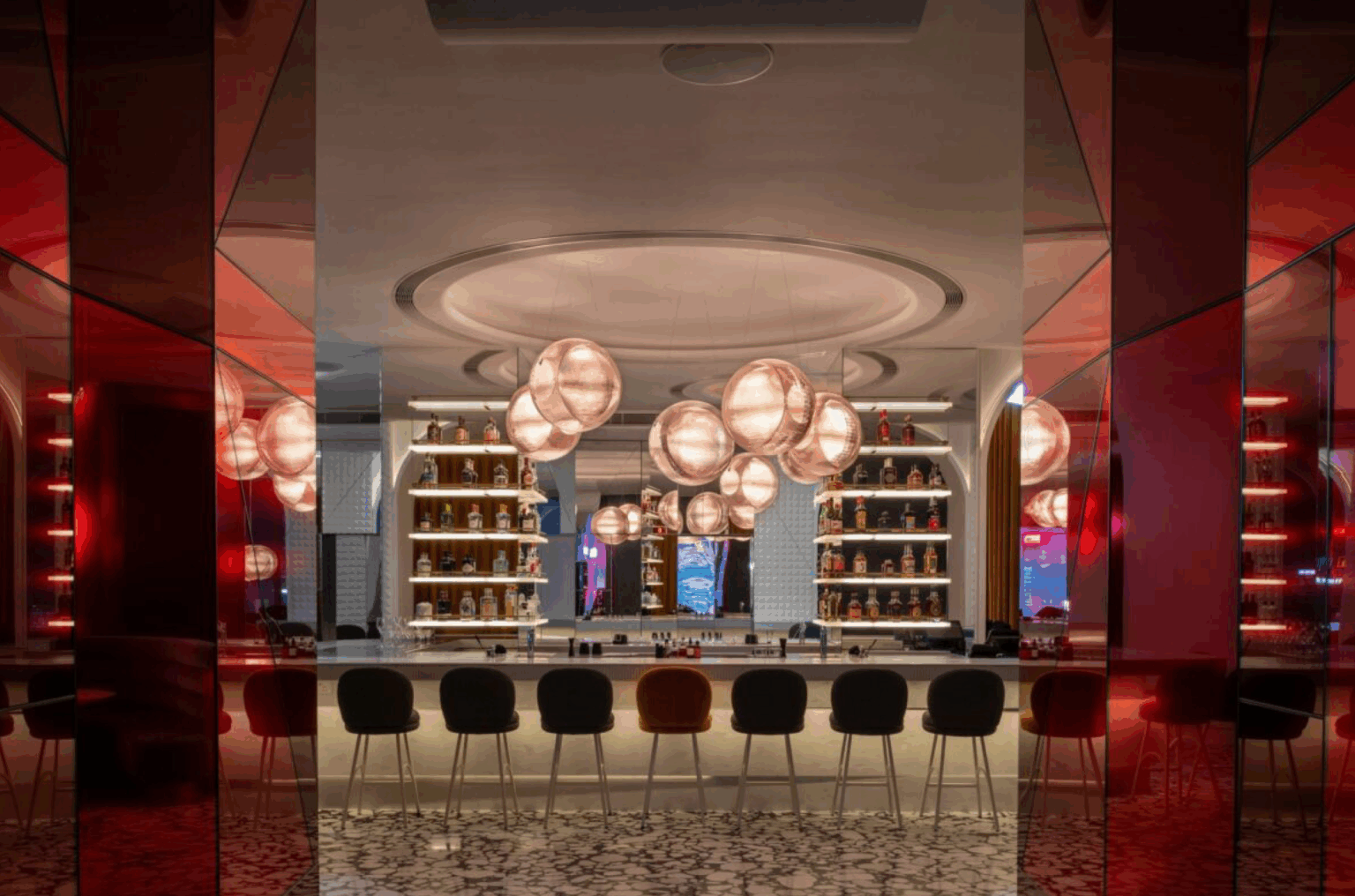 A modern hotel bar with a central circular counter, surrounded by high chairs. Soft, globe-like pendant lights hang above, illuminating shelves of bottles, creating a warm, inviting ambiance with red and gold tones.