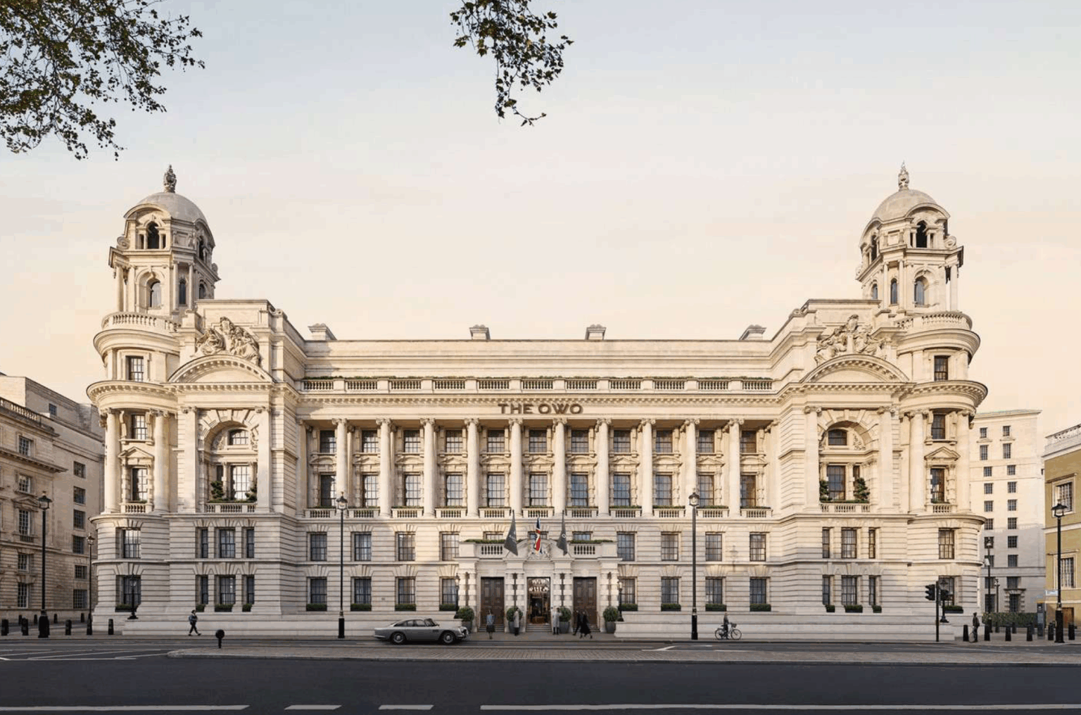 The OWO Residences by Raffles exteriors. Majestic neoclassical building with grand columns, ornate facades, and two towers; the setting is serene with a pale sky and sparse traffic.