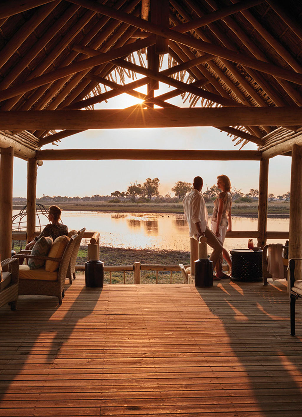 Rustic lodge interior with wooden furniture overlooks a serene waterway at sunset. Warm light casts shadows as two people stand by the water, creating a tranquil atmosphere.