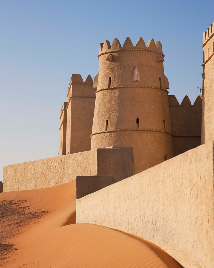 Ancient desert fortress with sand-colored walls and round towers under a clear blue sky. The foreground features red sand dunes creating a serene atmosphere.