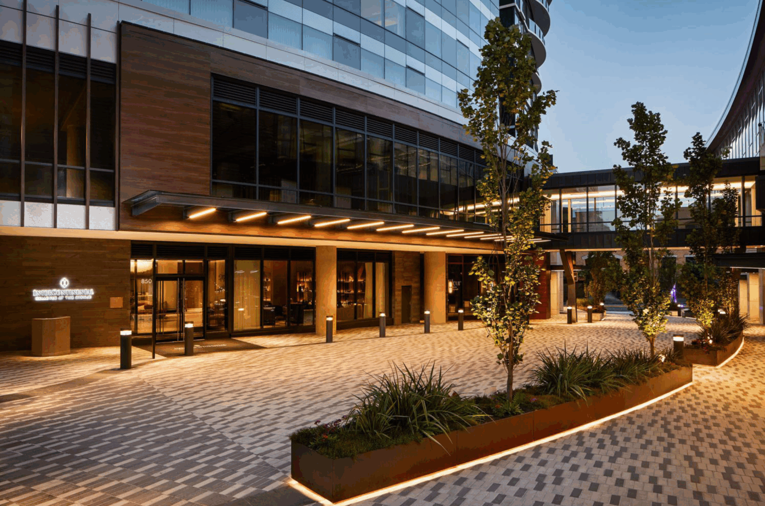 Modern hotel exterior at dusk with illuminated entrance, large windows, and trees in planters. Elegant and inviting atmosphere.