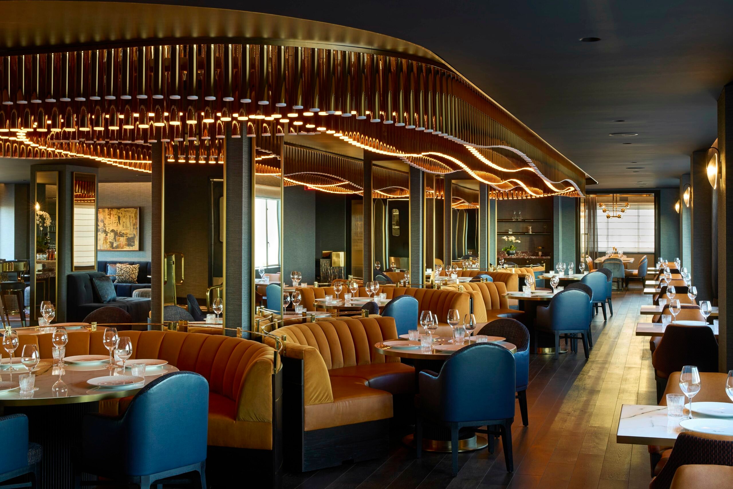 Luxurious hotel bar interior with warm lighting, featuring plush orange booths, blue chairs, elegantly set tables, and an undulating, golden ceiling fixture.
