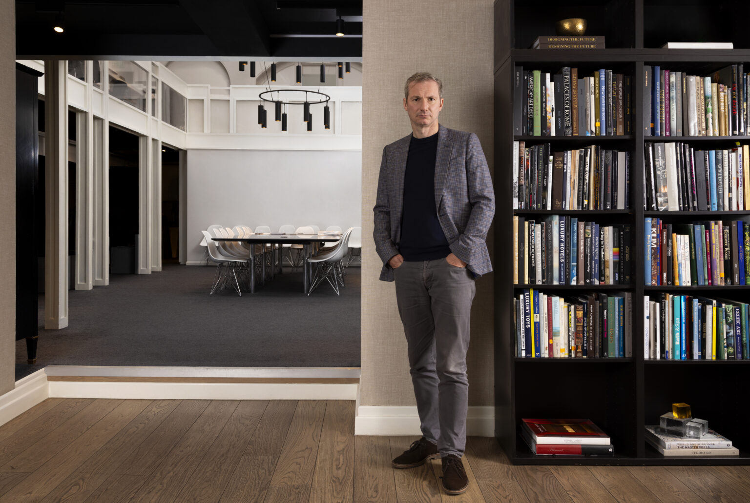 Portrait of HBA Co-CEO Chris Godfrey in a blue checked blazer standing near a bookcase filled with books. To the left, a modern dining room with white chairs and a black chandelier.