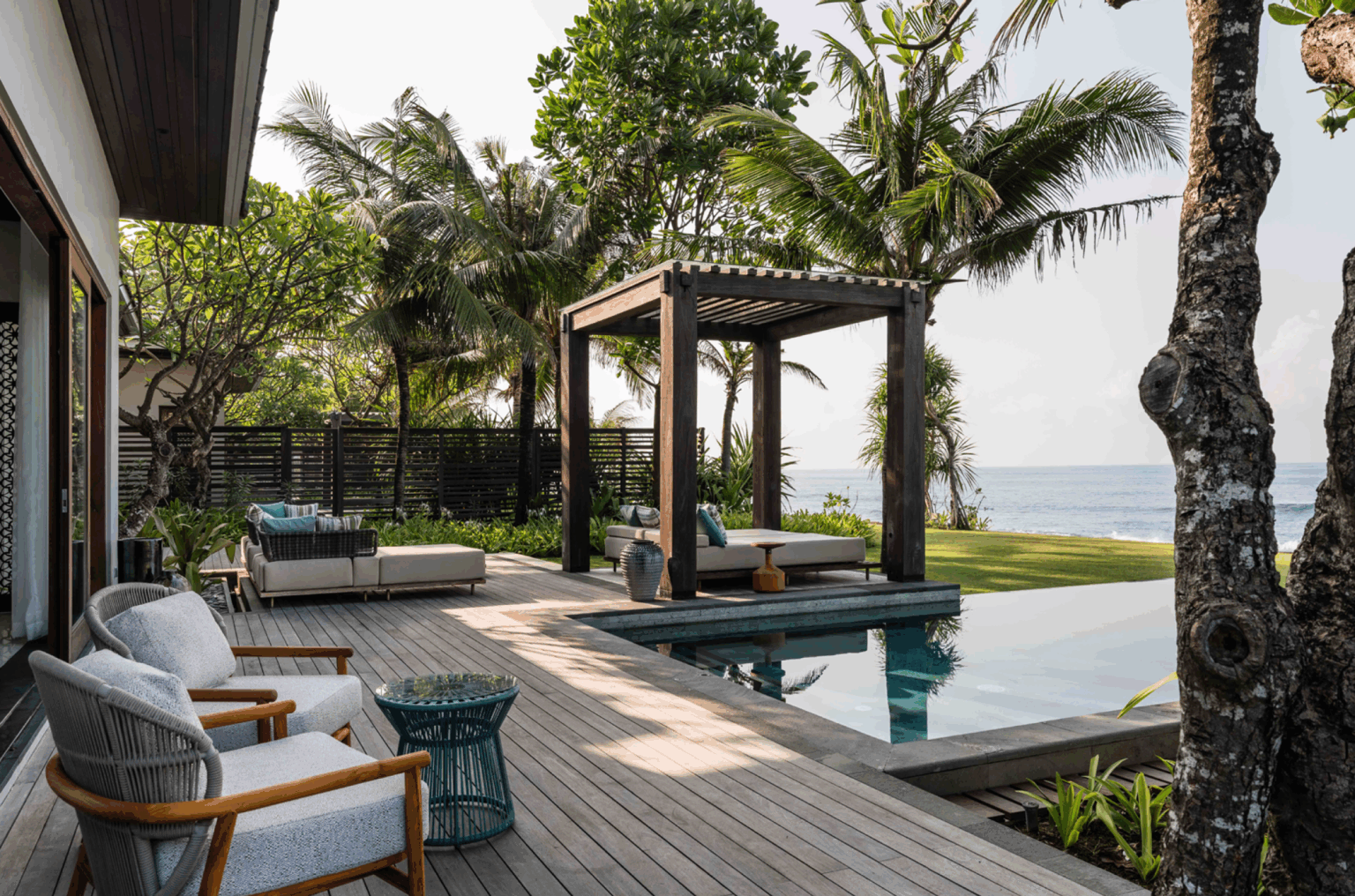Tropical poolside oasis with wooden deck, lounge chairs, and a pergola. Palm trees and ocean view create a serene, relaxing atmosphere.