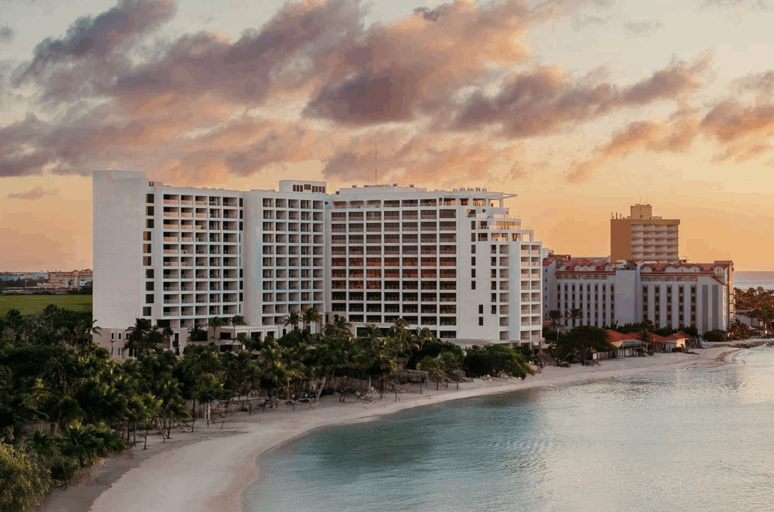 The St. Regis Aruba's exterior featuring a beachfront hotel with white facade at sunset, surrounded by palm trees and calm turquoise water. The sky is painted with pink and orange clouds, conveying tranquility.