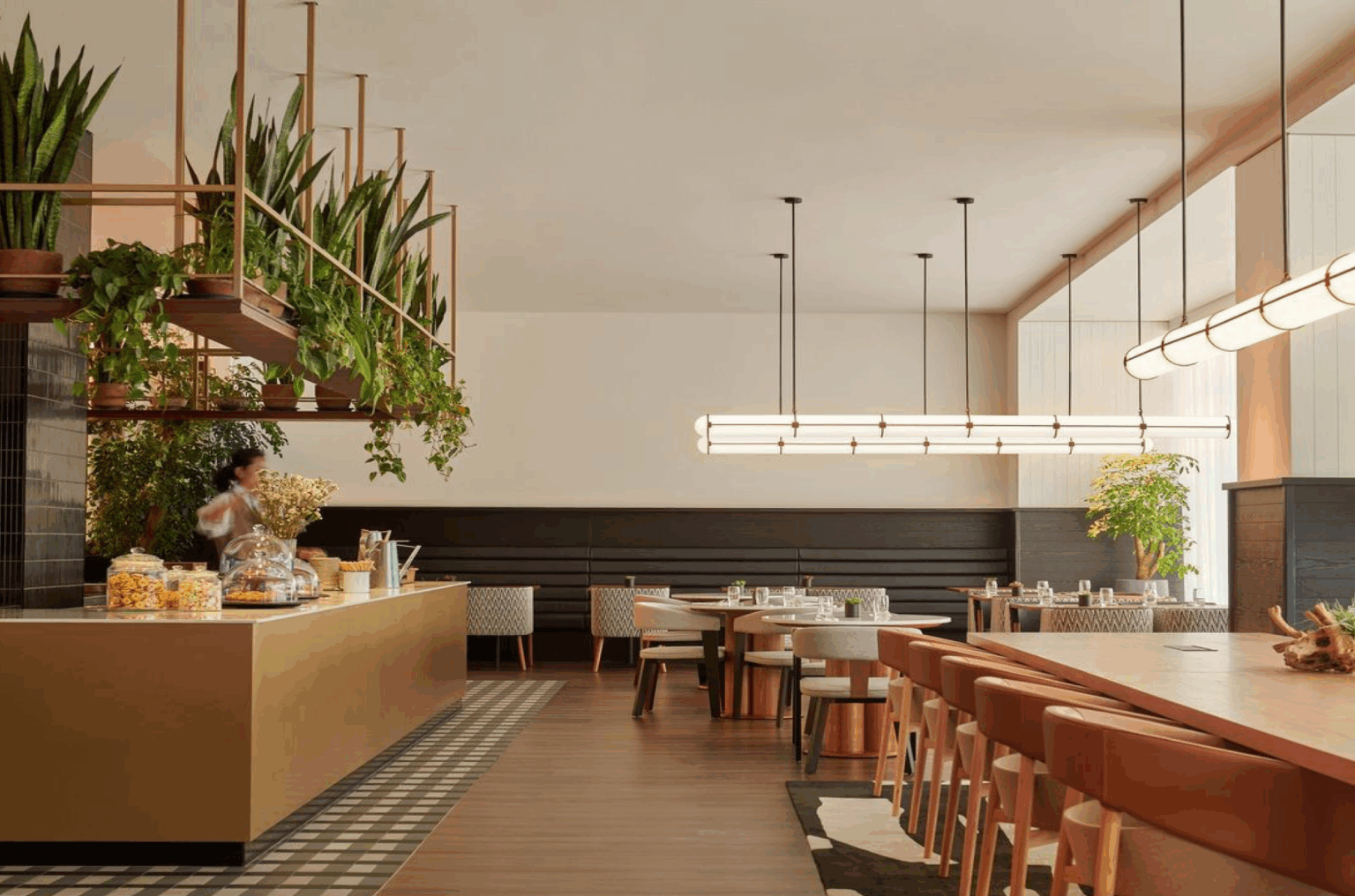 Modern hotel restaurant interior with a warm, inviting atmosphere. Suspended plants and sleek lighting hang over wooden tables and beige chairs. A person stands at a counter.