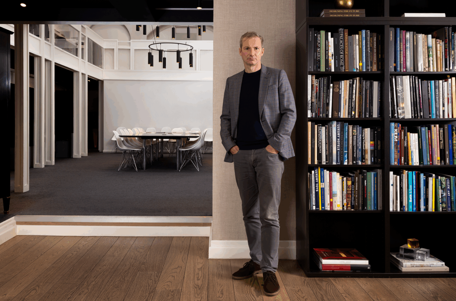 Portrait of HBA Co-CEO Chris Godfrey in a blue checked blazer standing near a bookcase filled with books. To the left, a modern dining room with white chairs and a black chandelier.