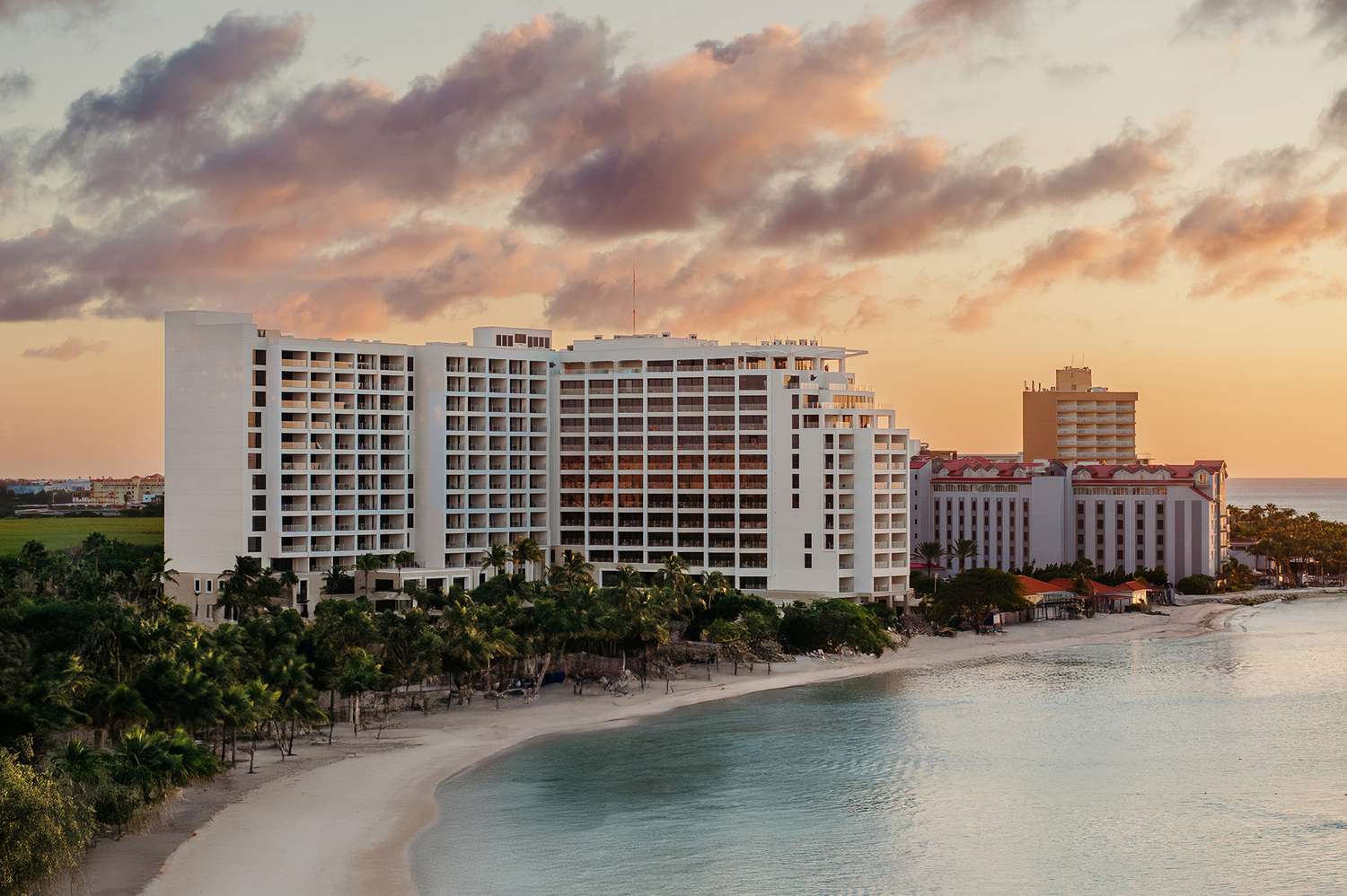 The St. Regis Aruba's exterior featuring a beachfront hotel with white facade at sunset, surrounded by palm trees and calm turquoise water. The sky is painted with pink and orange clouds, conveying tranquility.