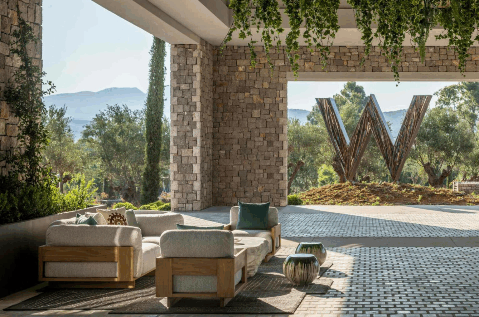 Outdoor hotel lounge area with wooden furniture and potted plants under a covered patio. Stone pillars frame a large 