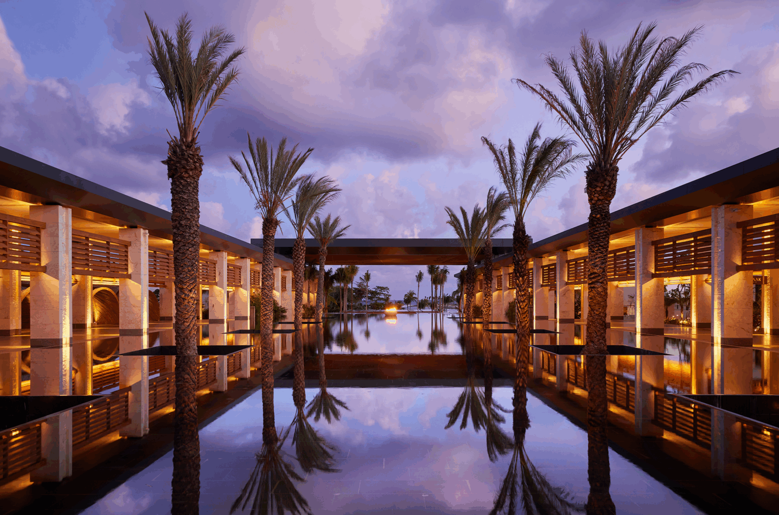 Twilight scene of a luxurious hotel courtyard with palm trees and warm lighting reflecting in a large, tranquil pool, creating a serene and elegant atmosphere.