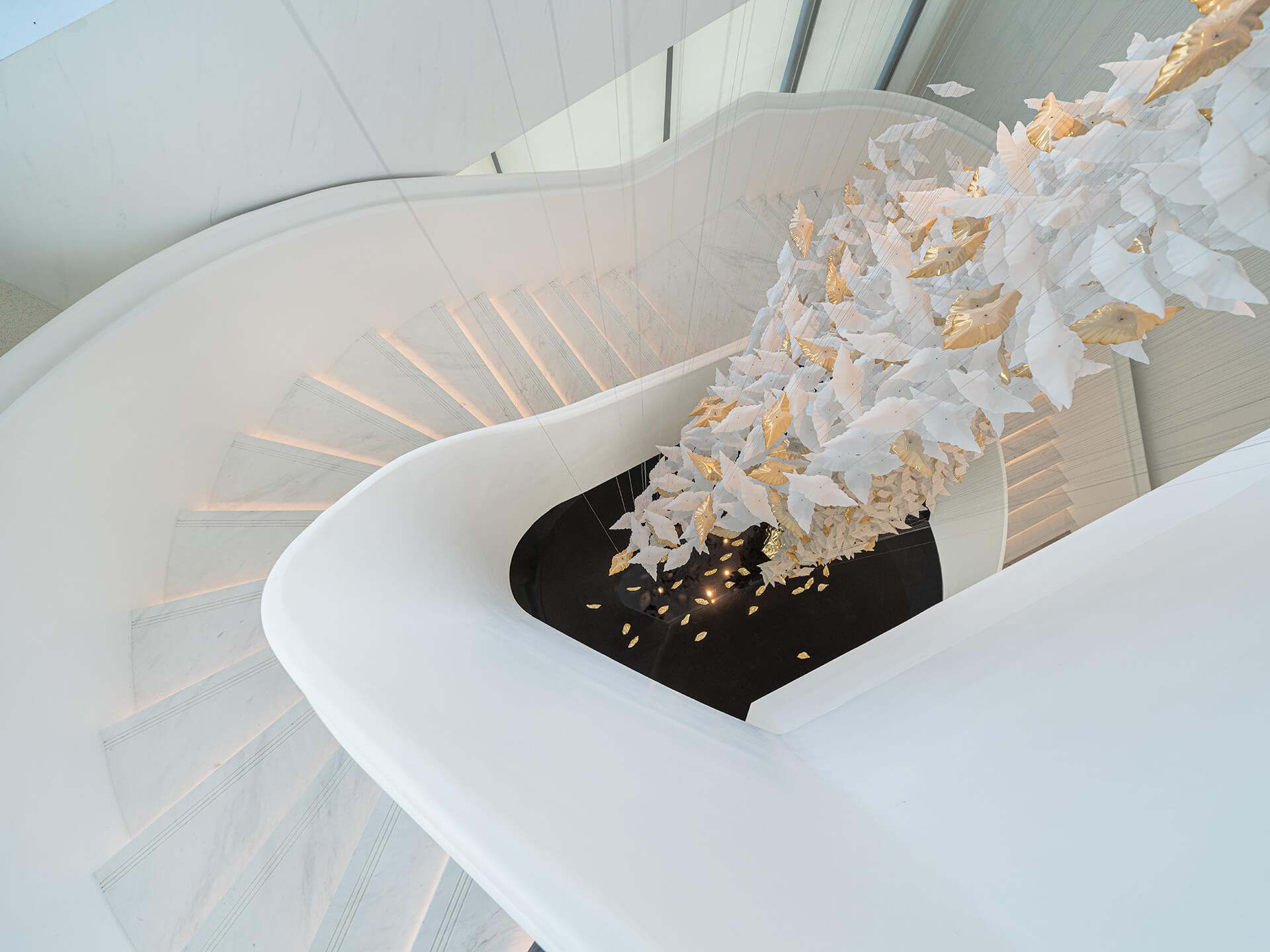 A white spiral staircase with subtle lighting descends around a large hanging art piece of white and gold leaves, creating an elegant, airy feel.