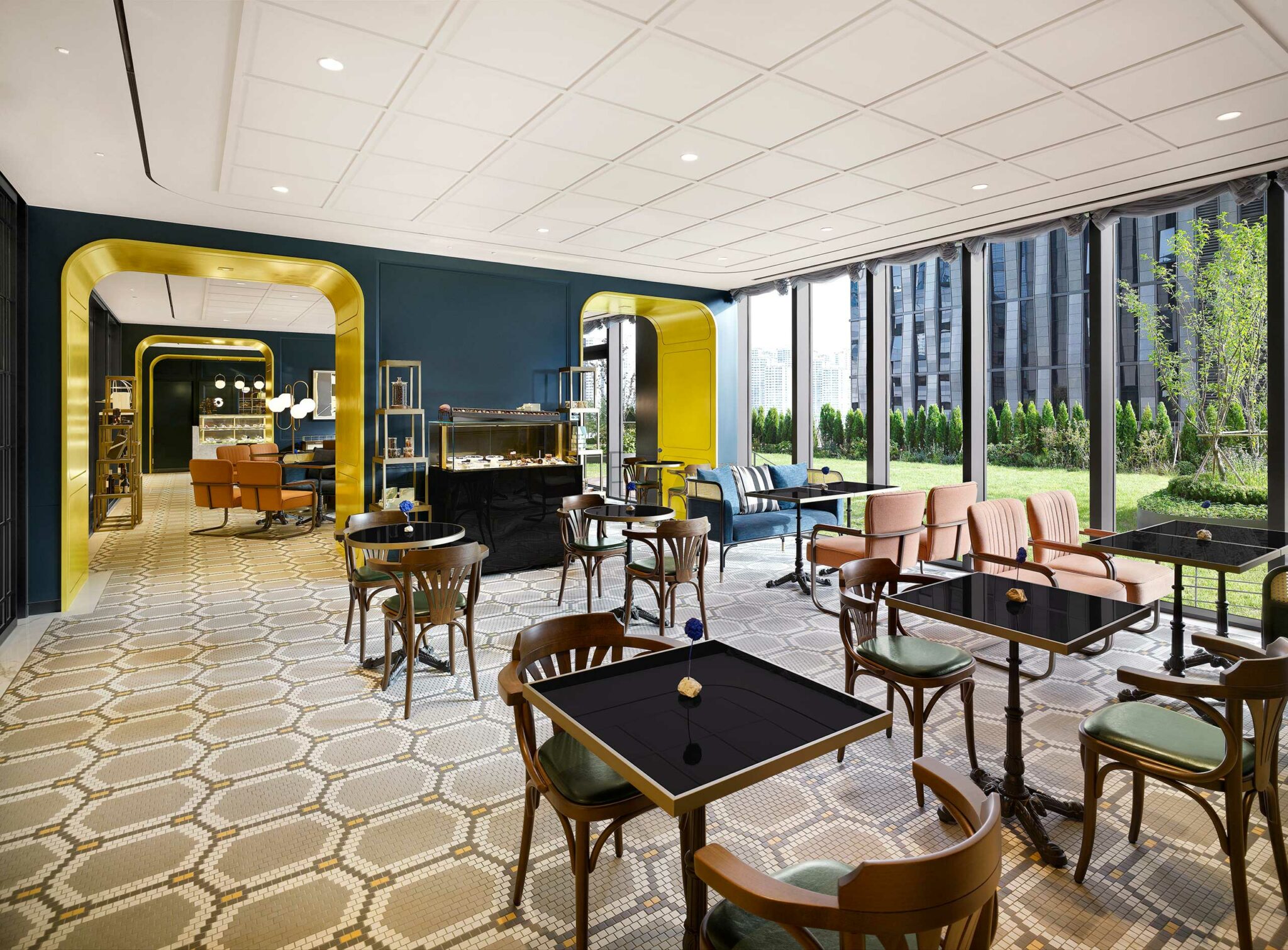 Modern hotel cafe with elegant decor, featuring dark blue walls, yellow arches, and patterned flooring. It has wooden tables, plush chairs, and large windows overlooking greenery, creating an inviting and stylish atmosphere.