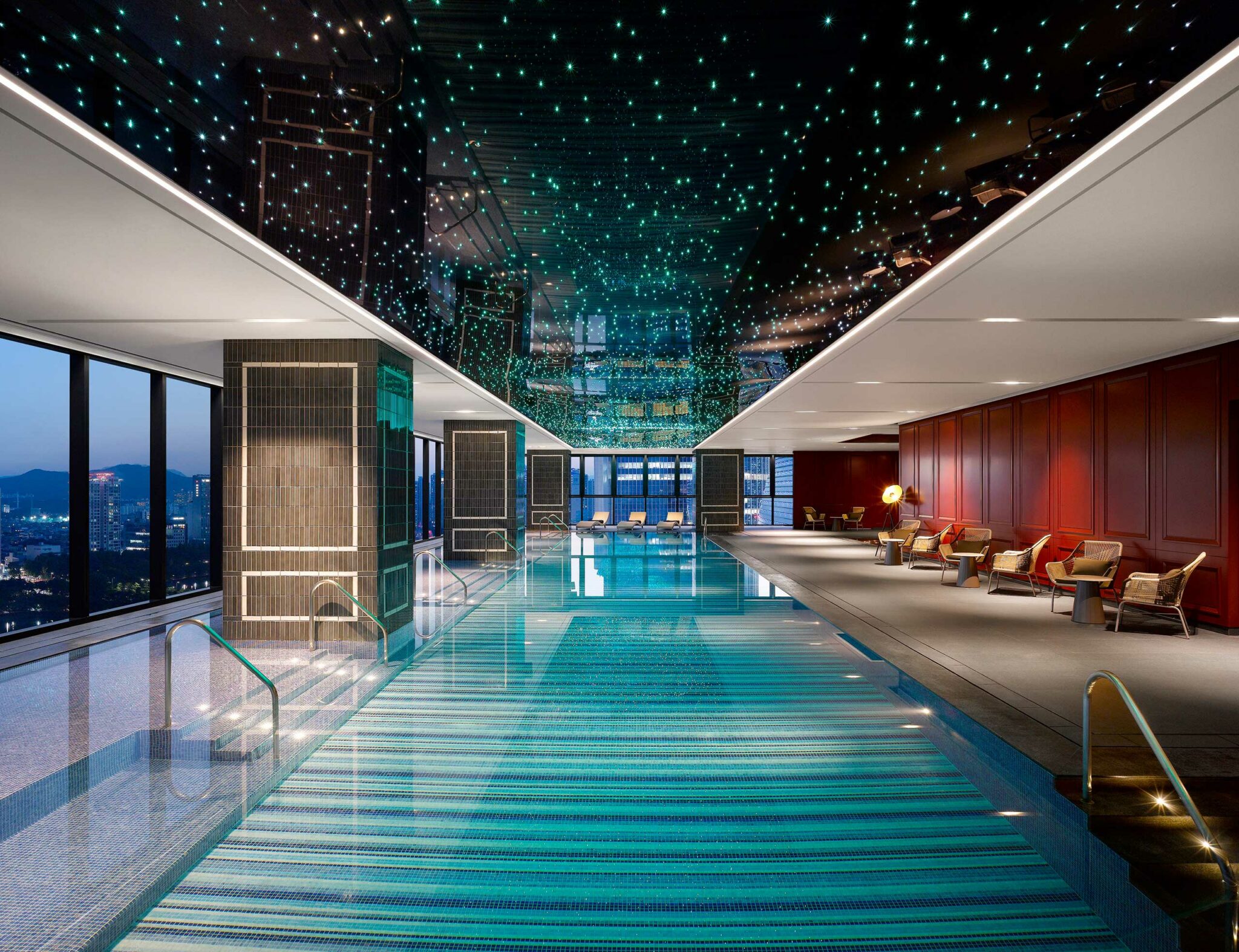 Luxurious hotel indoor pool with a starry ceiling, surrounded by modern lounge chairs. Large windows on the left offer a nighttime city view. Tranquil ambiance.