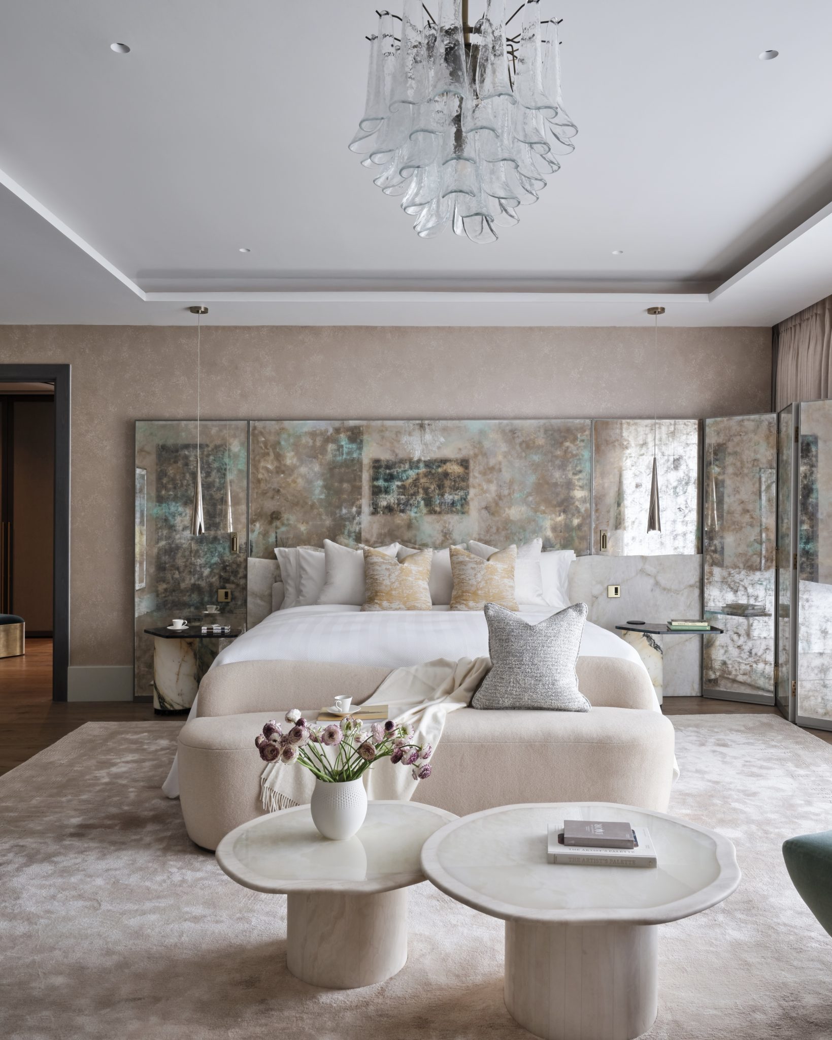 Luxurious hotel room with a king-sized bed, elegant decor, and large windows offering a stunning city view. Pastel tones and plush seating create a cozy, sophisticated ambiance.