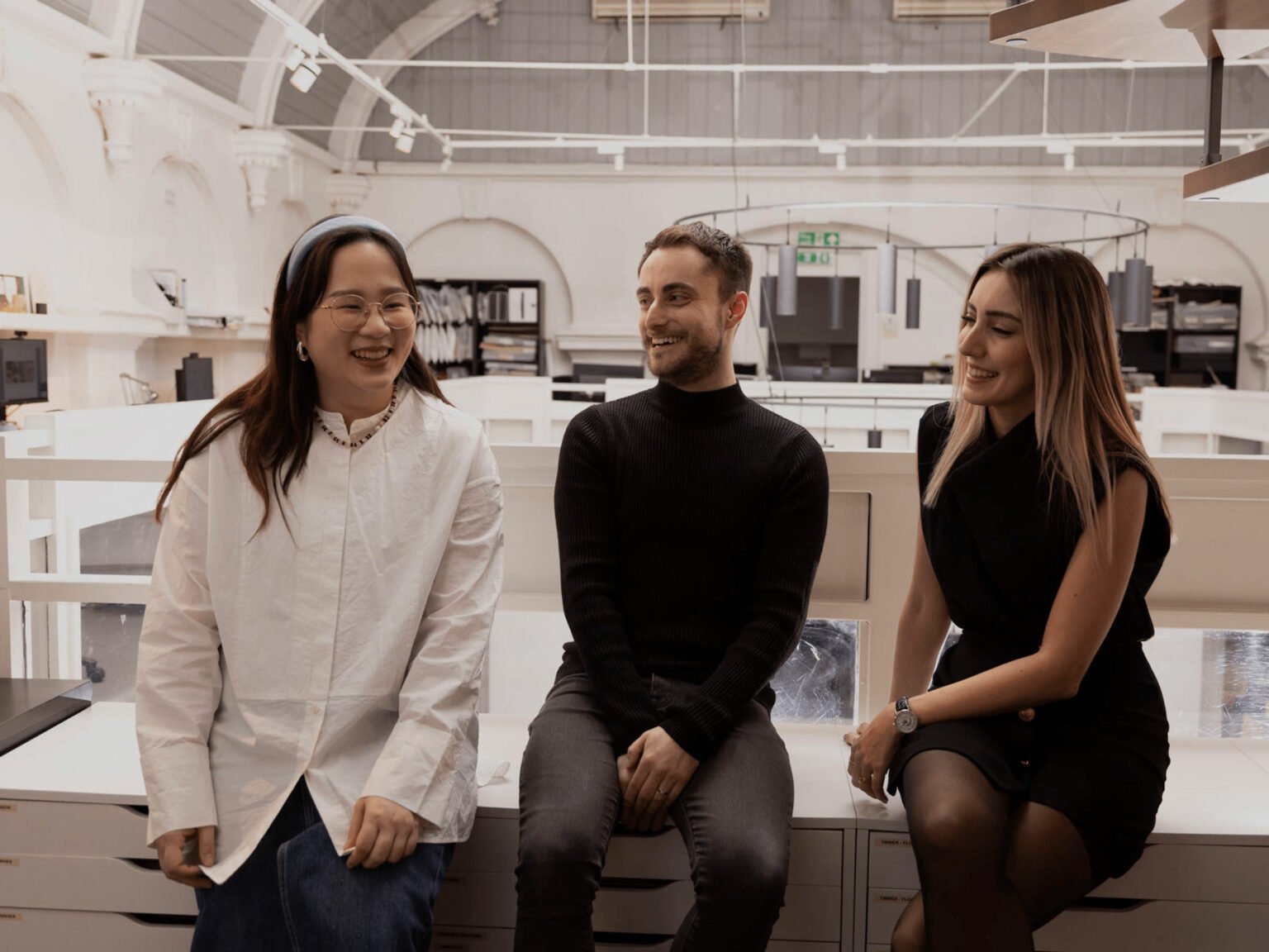 Three young designers sitting on desks in a bright, modern office setting, smiling and engaged in a casual conversation, conveying a friendly atmosphere.
