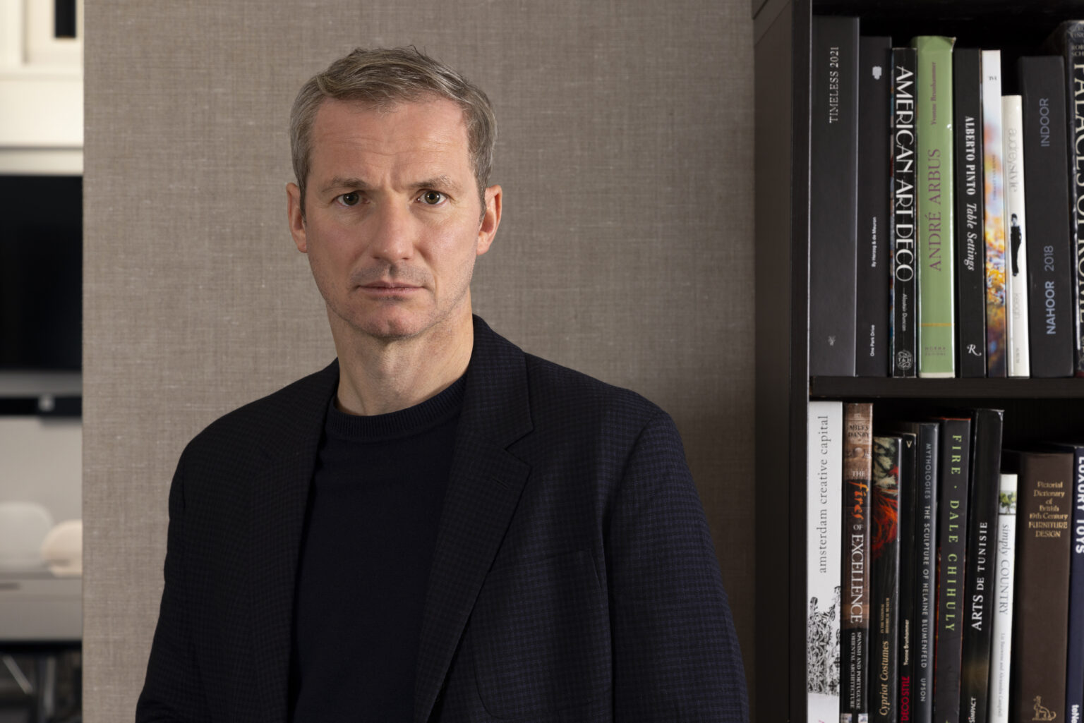 Portrait of HBA Co-CEO Chris Godfrey wearing a dark blazer and sweater standing against a textured beige backdrop, looking serious and contemplative.