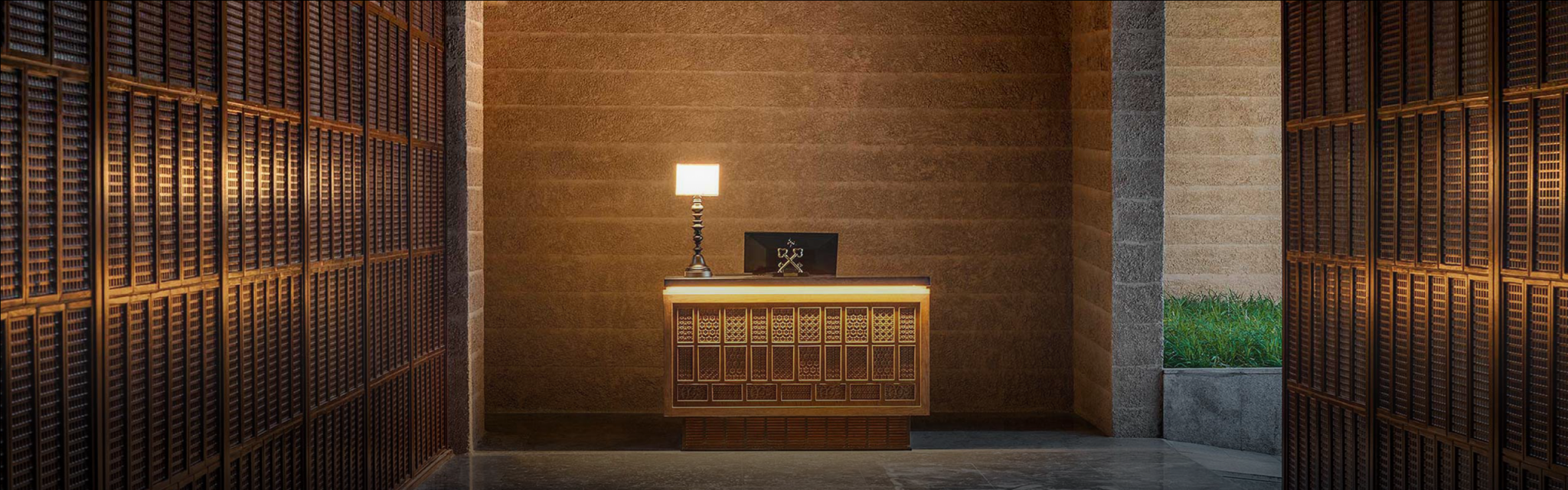 Hotel reception area with warm lighting, featuring a wooden desk and table lamp against a textured wall. Flanked by lattice-patterned panels, creating an inviting atmosphere.