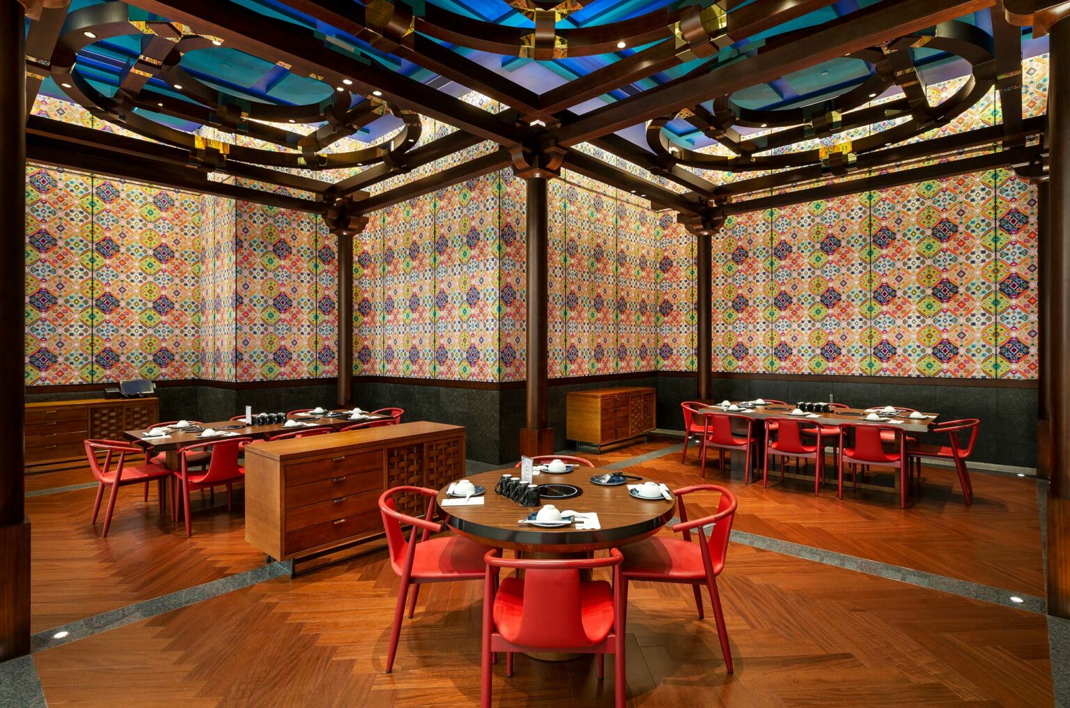 Sophisticated hotel dining area with round tables, red chairs, and colorful mosaic walls. The warm wood flooring and artistic ceiling add elegance.