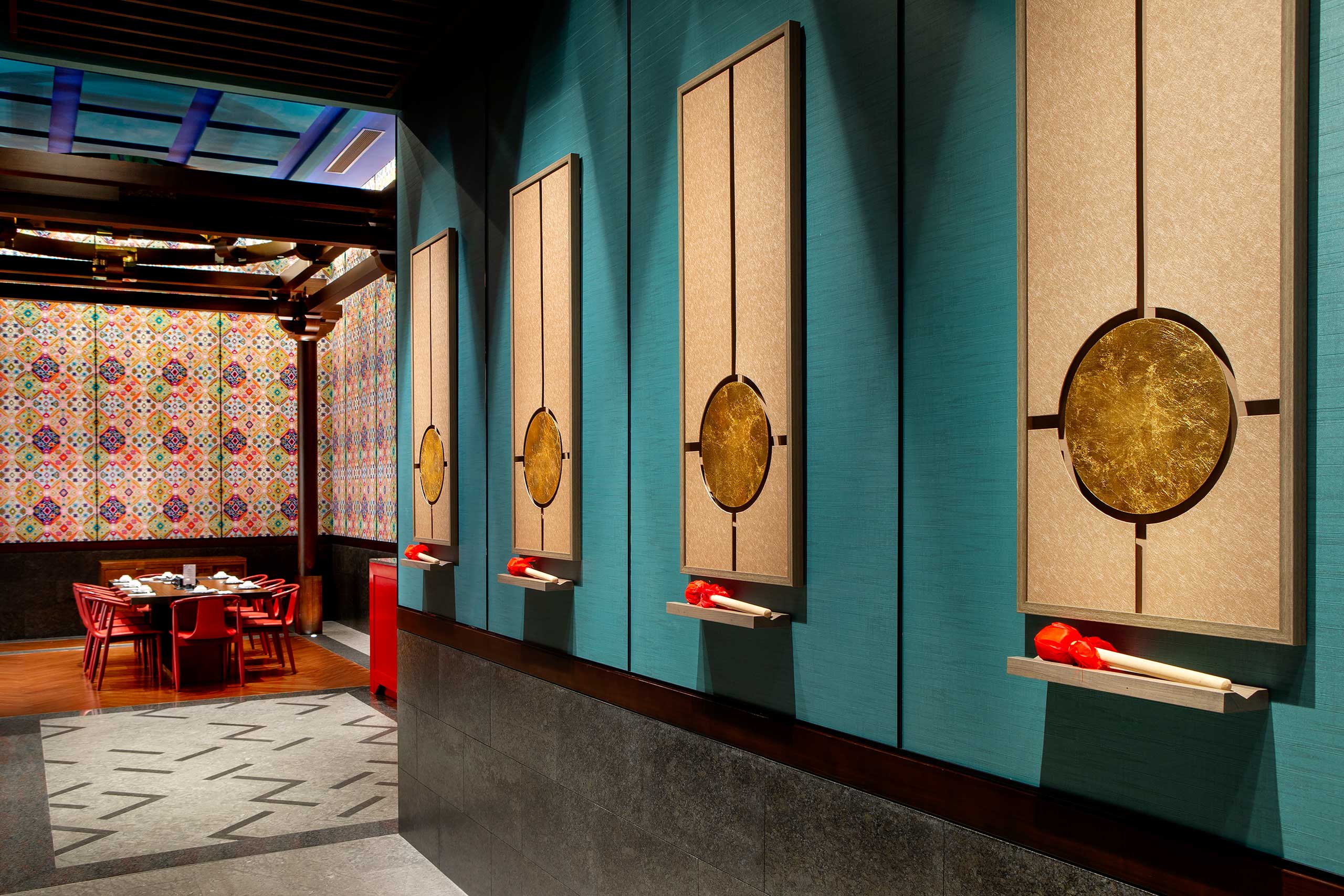 Modern hotel restaurant interior with teal walls featuring abstract art panels and red decorative items. In the background, a dining area with a patterned wall and red chairs creates a vibrant, stylish ambiance.