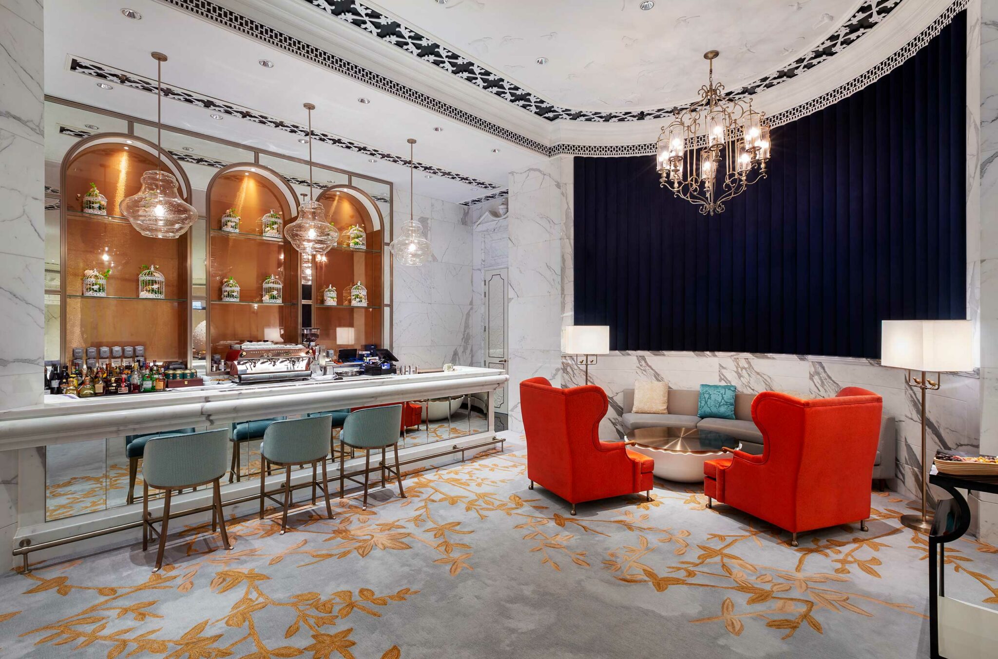 Elegant hotel lounge with a marble countertop bar, teal barstools, hanging pendant lights, vibrant red armchairs, and a chandelier, exuding luxury.
