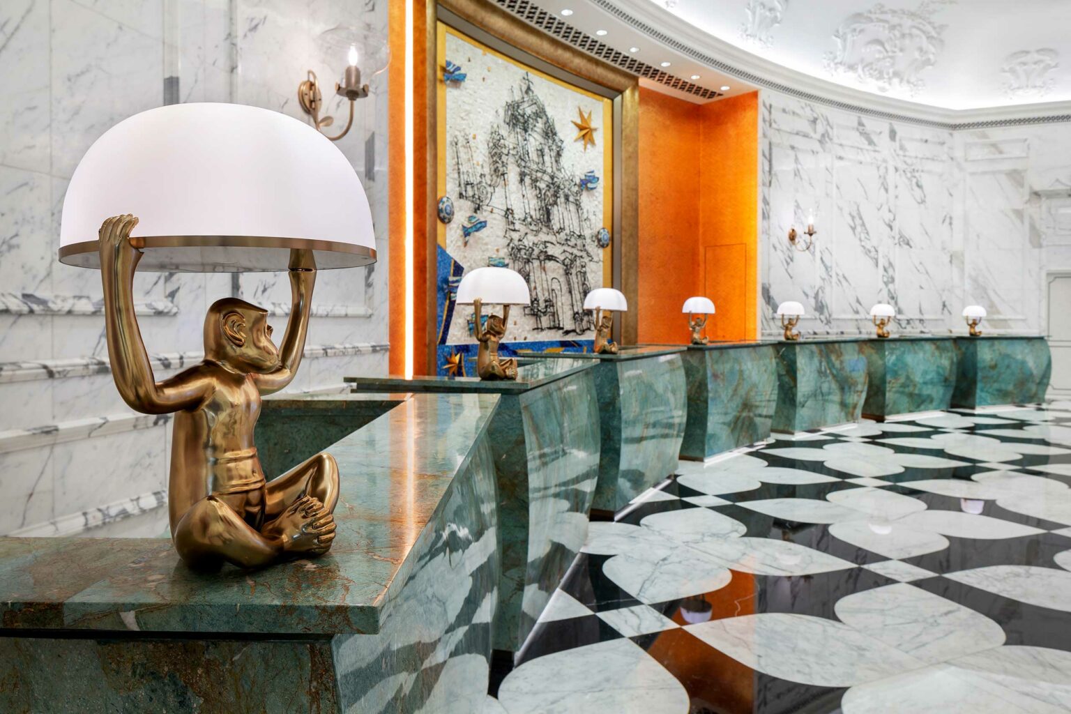 Luxurious marble hotel lobby with elegant décor. Brass monkey lamps holding wide dome lights sit atop a long, curved, green marble reception desk. Walls adorned with intricate reliefs and a large mosaic artwork in the background create a refined atmosphere.