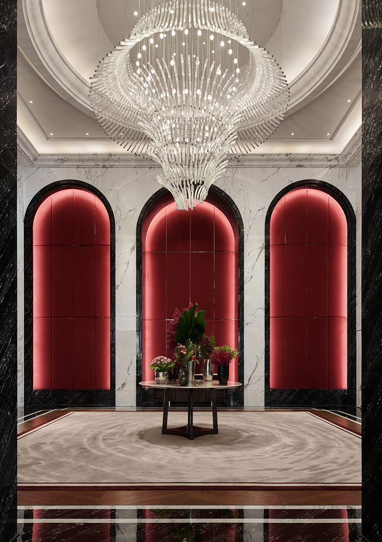 A luxurious hotel lobby featuring a grand chandelier, elegant arched design elements, and rich red accents, complemented by a central table adorned with flowers, creating a sophisticated and welcoming ambiance.