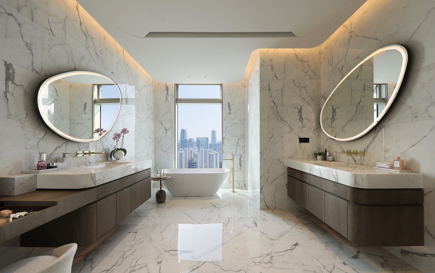 Luxurious marble bathroom with two oval mirrors, a central freestanding tub, and a cityscape view. Warm lighting creates an elegant atmosphere.