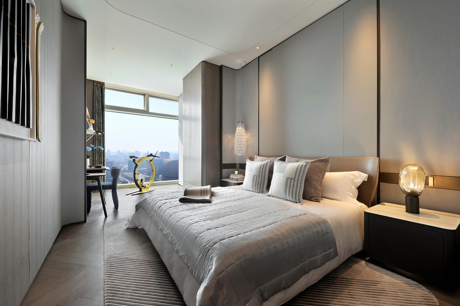 Modern bedroom with a cozy beige bed, two plaid pillows, and a plush bench at the foot. Sunlight streams through a large window, enhancing warmth.