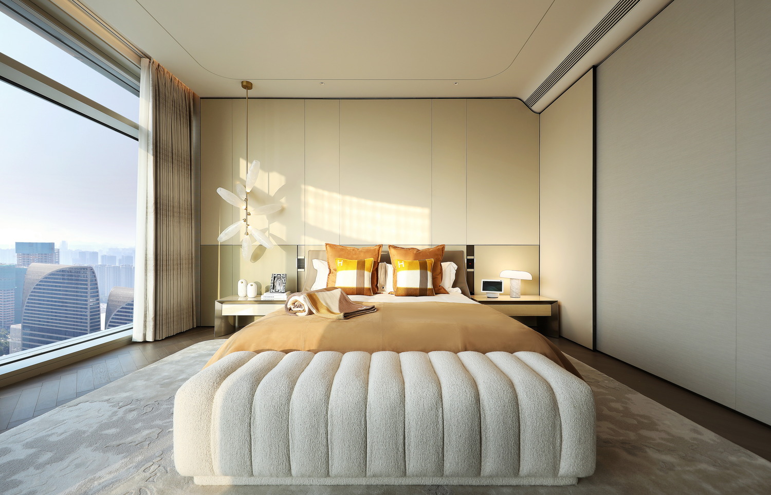 Modern bedroom with a cozy beige bed, two plaid pillows, and a plush bench at the foot. Sunlight streams through a large window, enhancing warmth.