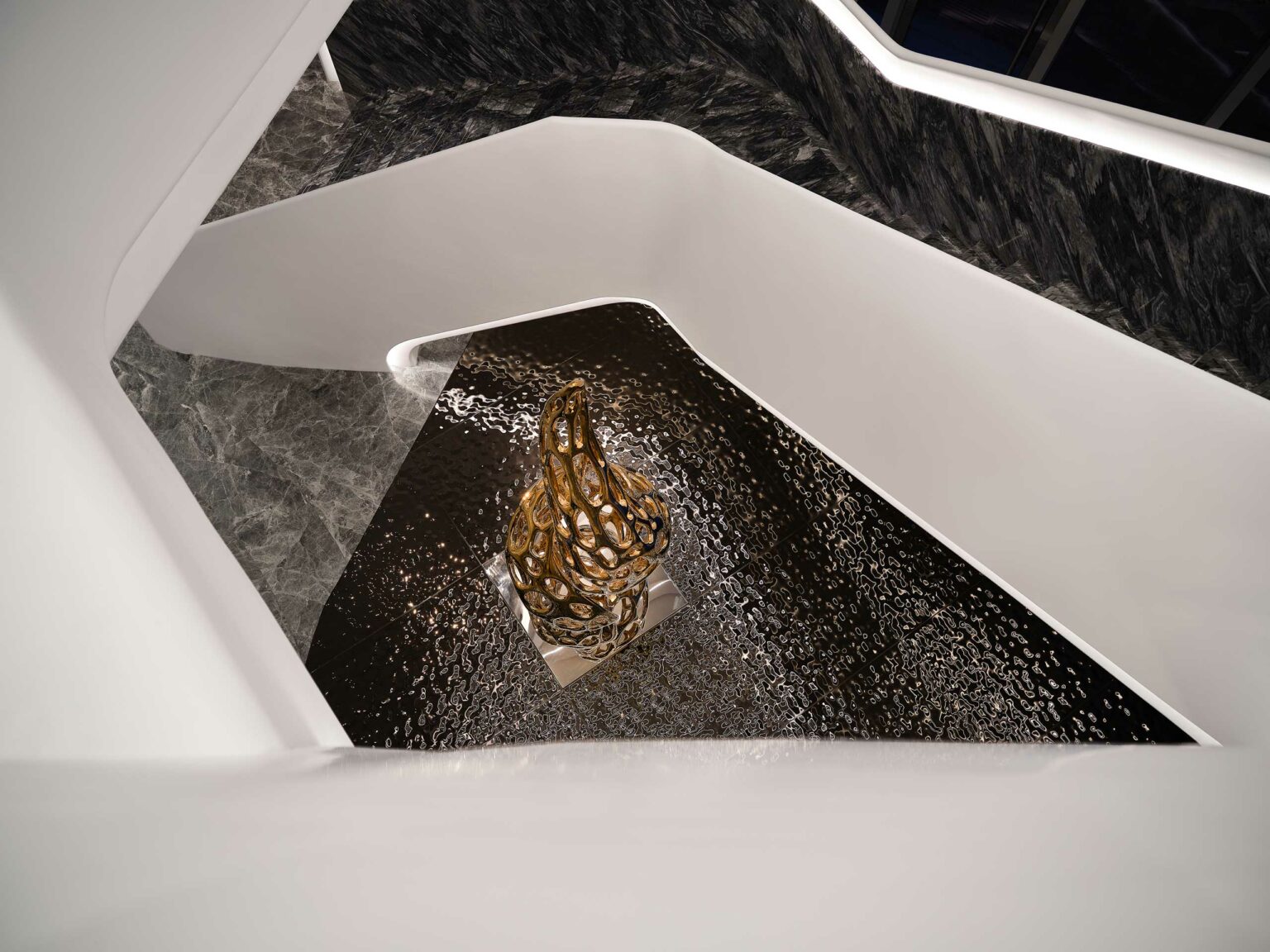 Overhead view of a modern sculpture in a spiraling design, set on a reflective, textured black surface, surrounded by sleek white and gray walls.