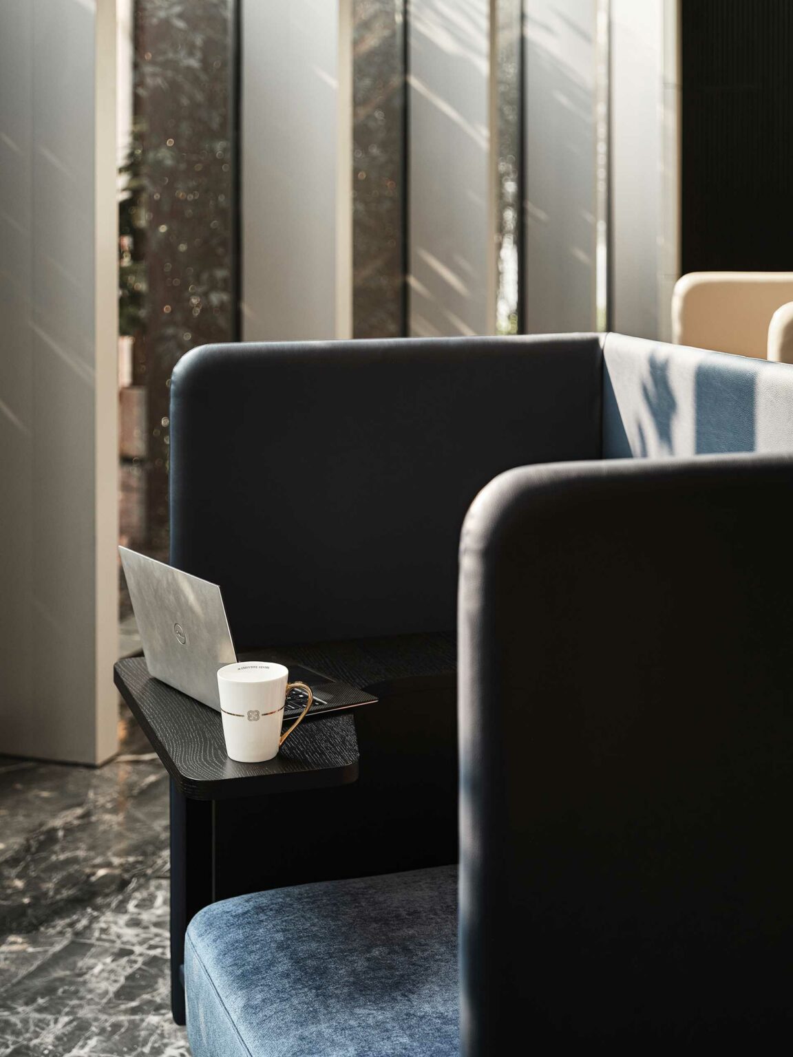 A modern workspace with a cozy booth features a laptop, a coffee cup, and glasses on a table. Soft sunlight streams through large windows, creating a serene atmosphere.