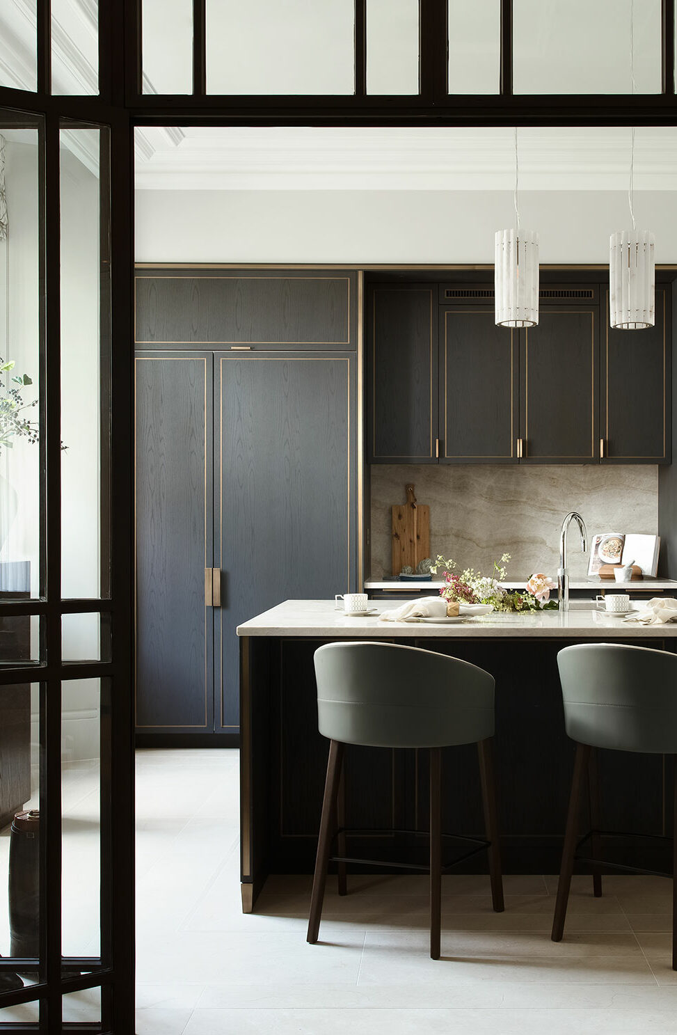 Sleek kitchen interior with dark cabinets and brass accents. Marble island with two green chairs, pendant lights, and a vase with flowers. Cozy and elegant.