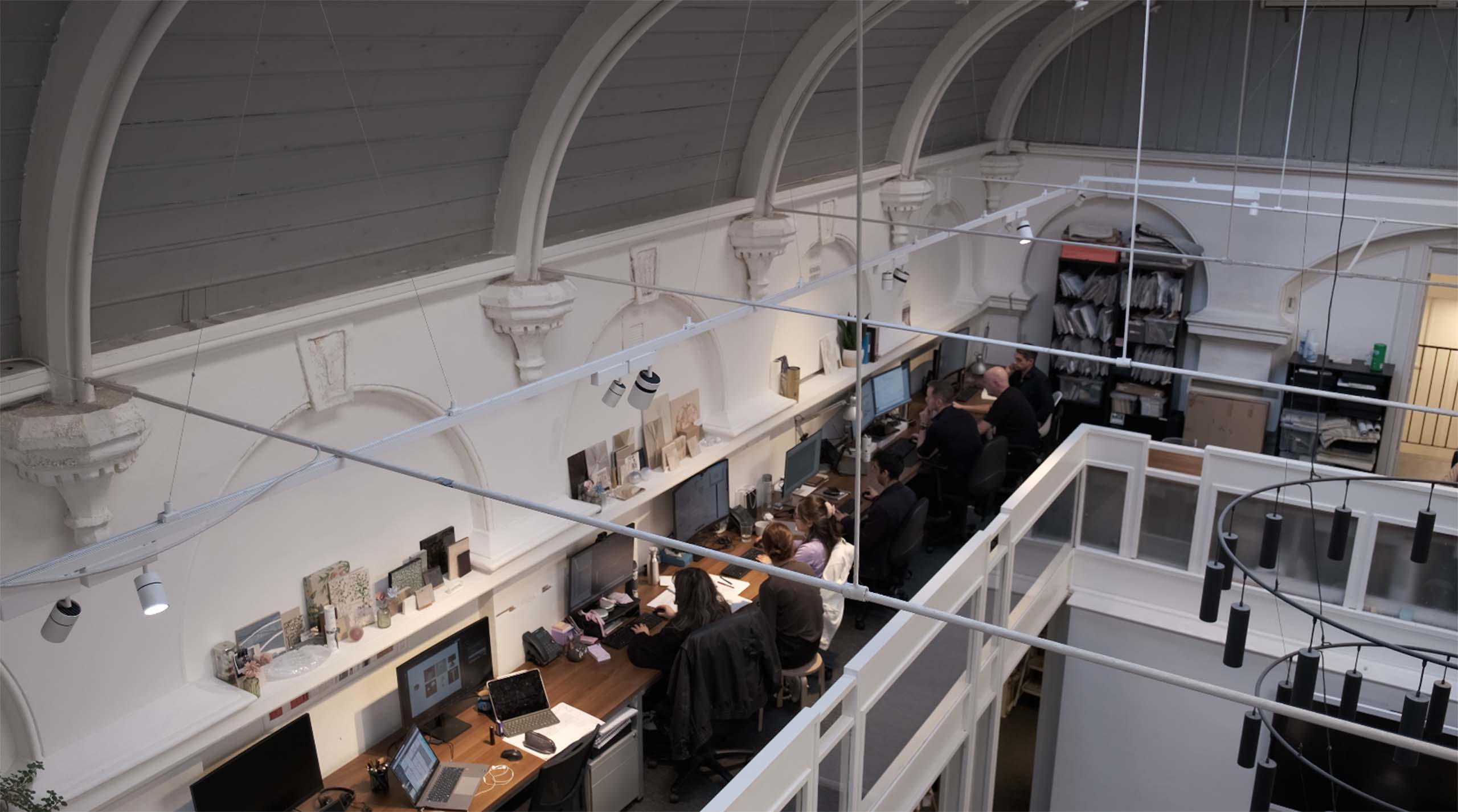 A modern design studio with an arched ceiling, featuring long desks with computers and four people working. The atmosphere is industrious and focused.