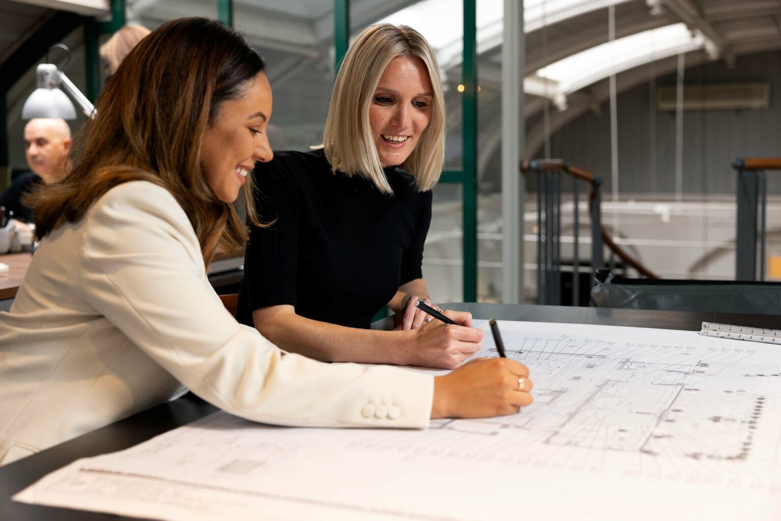 Two female designers smiling, collaborating on architectural blueprints at a desk in a modern office. The atmosphere is professional and positive.