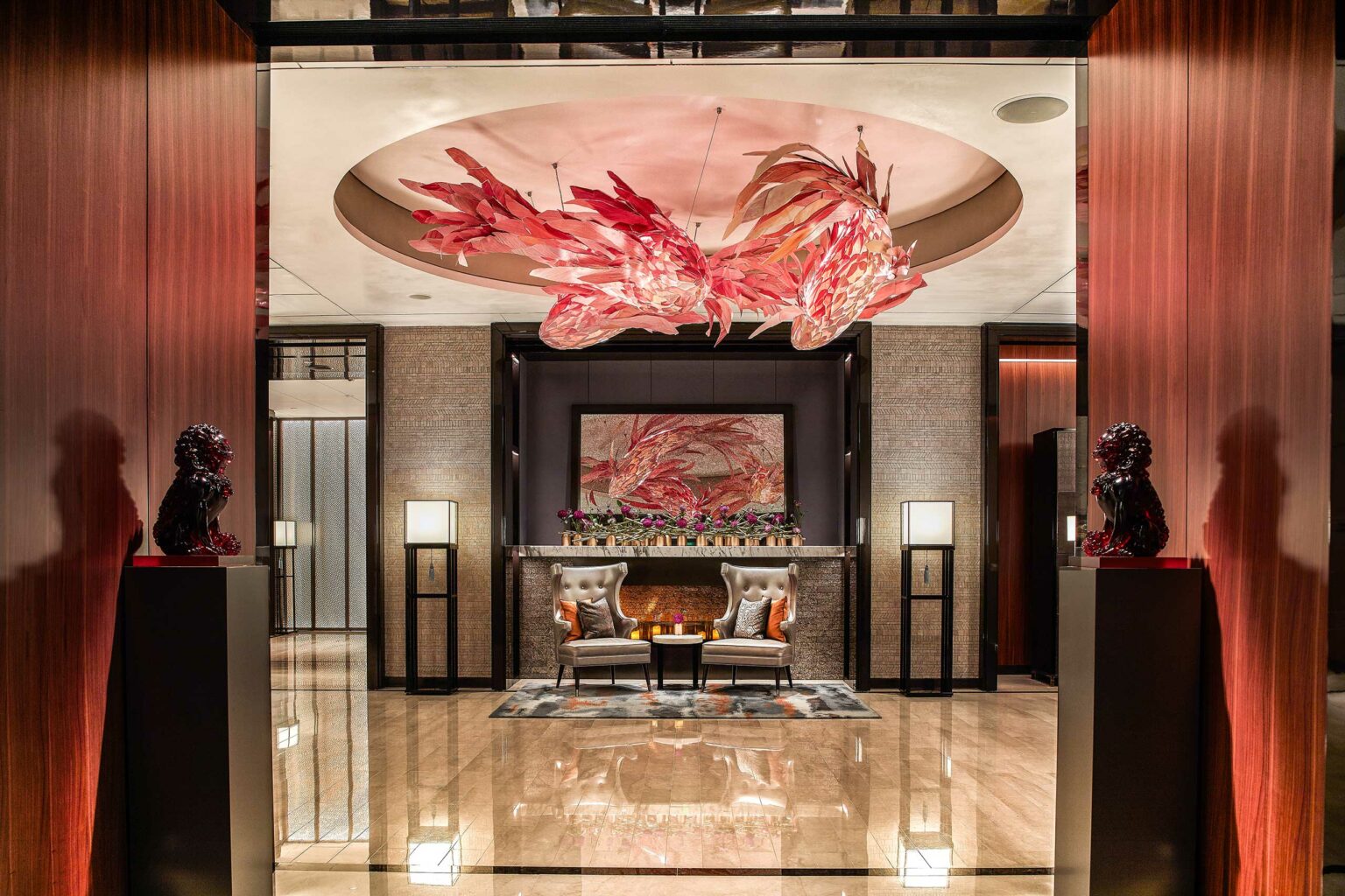 Luxurious hotel lobby with a red glass chandelier overhead, modern decor, two grey chairs, and art on the wall. Warm lighting creates an elegant atmosphere.