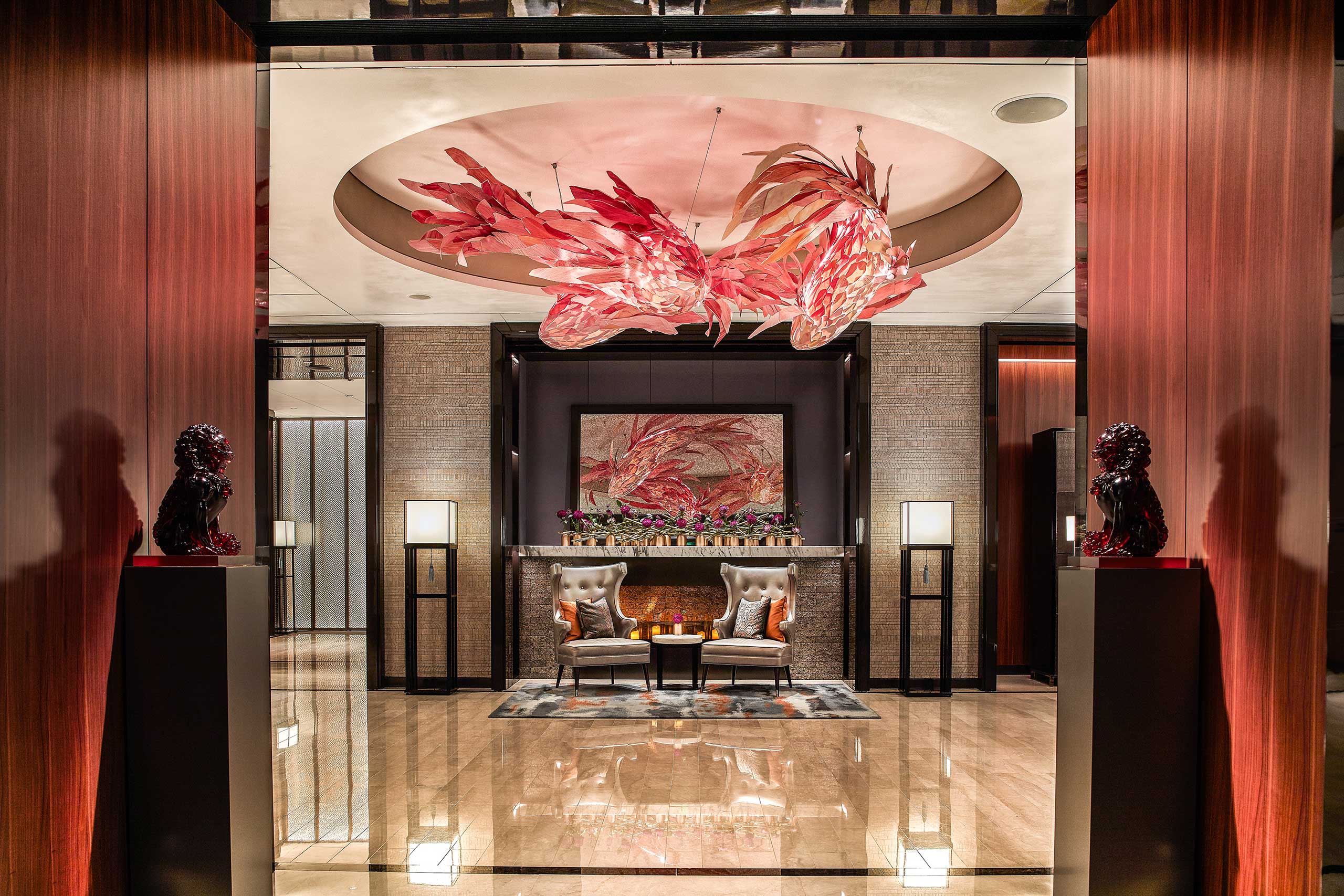 Luxurious hotel lobby with a red glass chandelier overhead, modern decor, two grey chairs, and art on the wall. Warm lighting creates an elegant atmosphere.