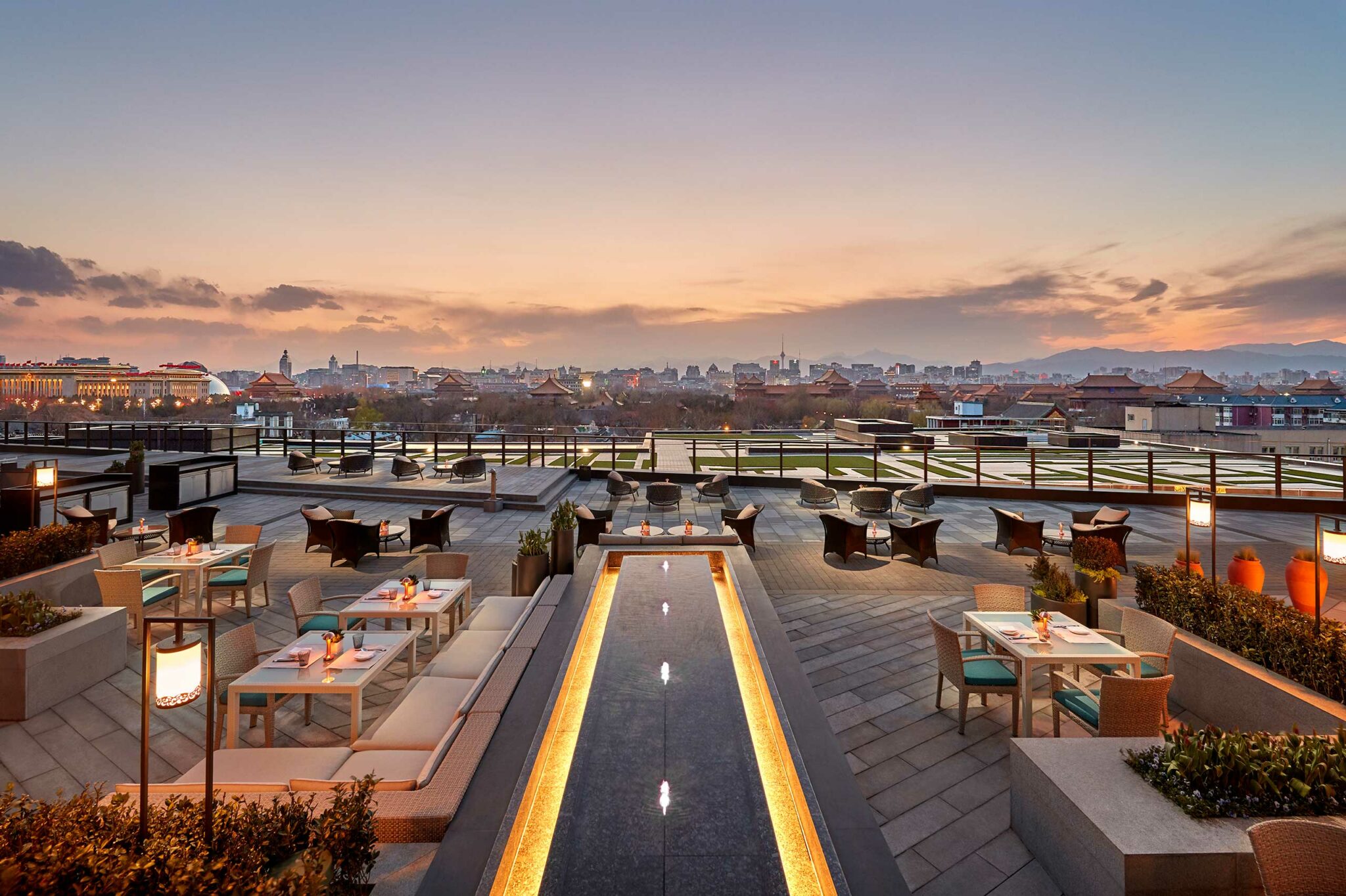 Hotel rooftop terrace at sunset with modern dining setup and water feature. Cityscape view, warm ambiance, and soft lighting create a tranquil atmosphere.
