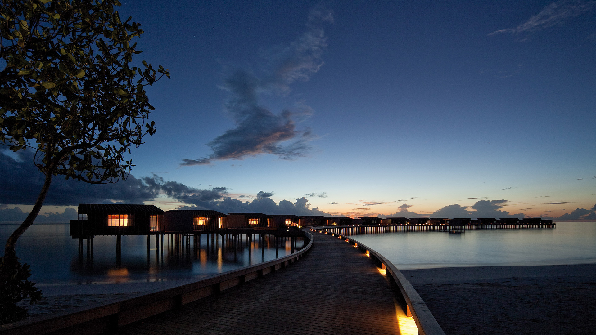 A serene dusk view of overwater bungalows with warm lighting, reflected on calm water. A curved wooden walkway leads toward a clear twilight sky.