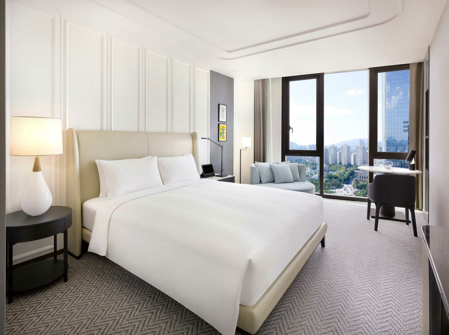 Modern hotel room with a large bed, white bedding, nightstand, and a lamp. Includes a sofa, desk, chair, and large windows with a city view.