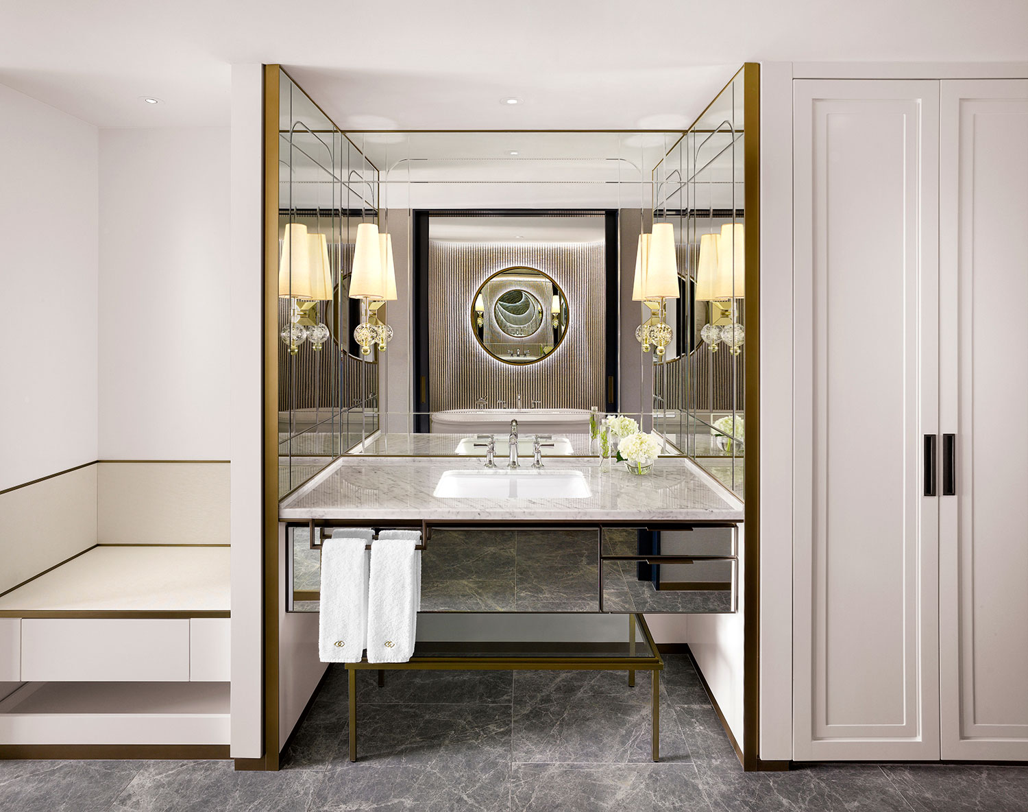 Luxurious suite bathroom with marble sink, ornate wall mirrors, gold accents, dual elegant lamps, and fresh flowers, exuding a serene, upscale ambiance.