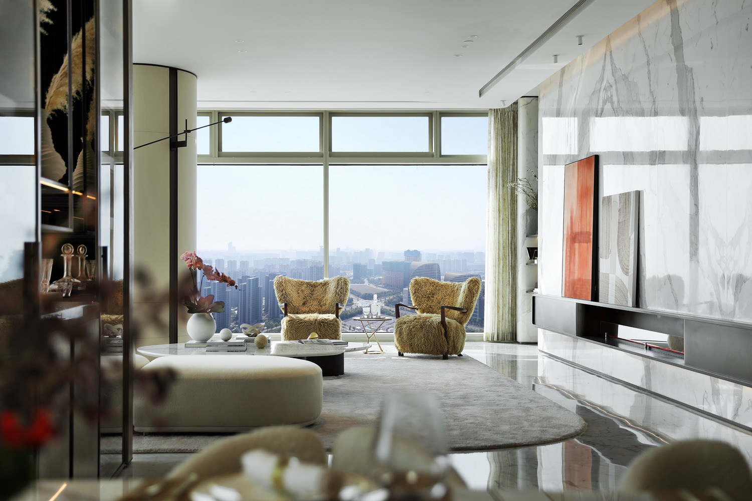 A modern living room with large windows, sunlight streaming in. Features include a plush chair, abstract wall art, round table, and curved sofa. The ambiance is serene and sophisticated.