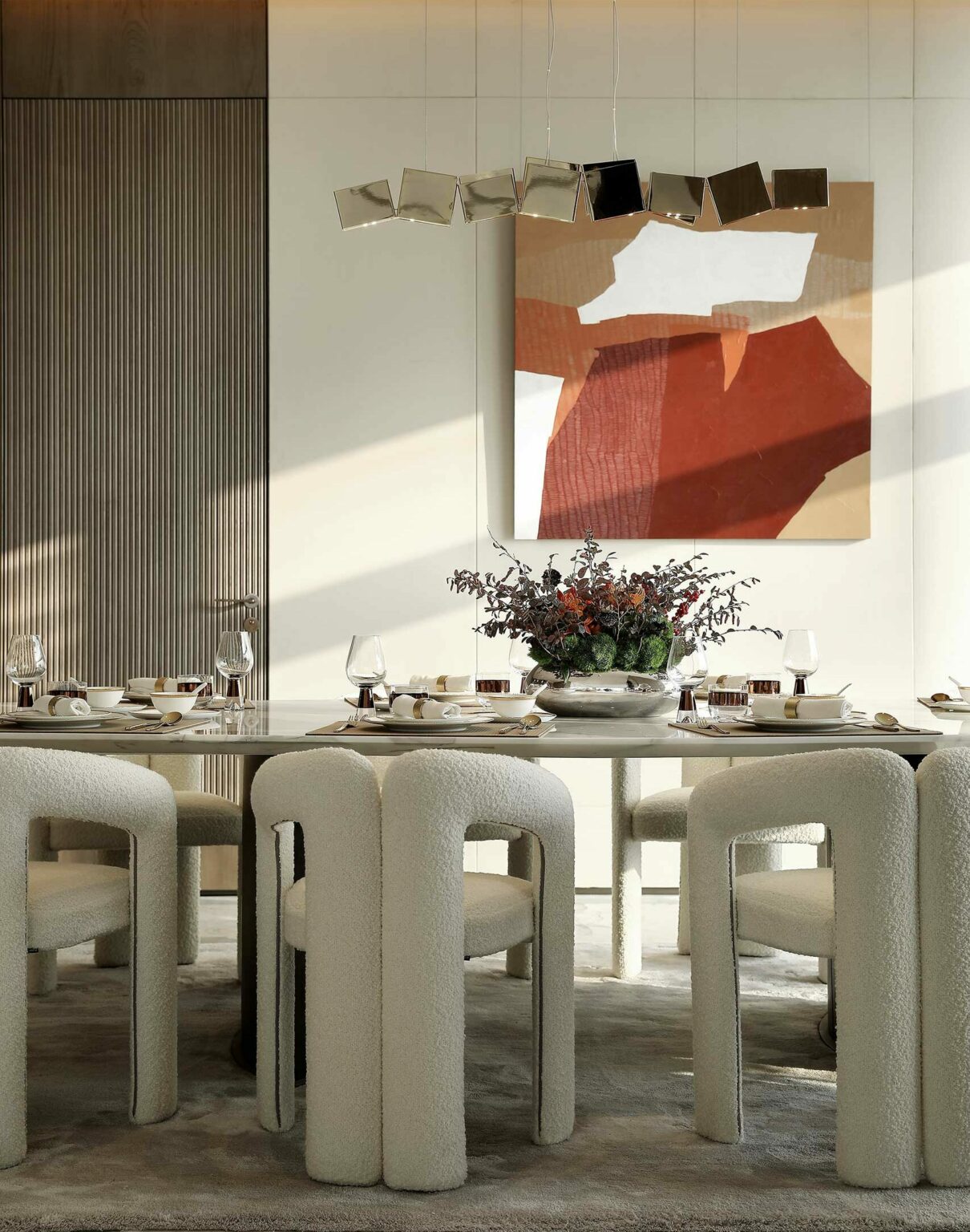 Modern dining room with a light marble table, white upholstered chairs, abstract red-brown artwork, metallic light fixture, and a floral centerpiece.