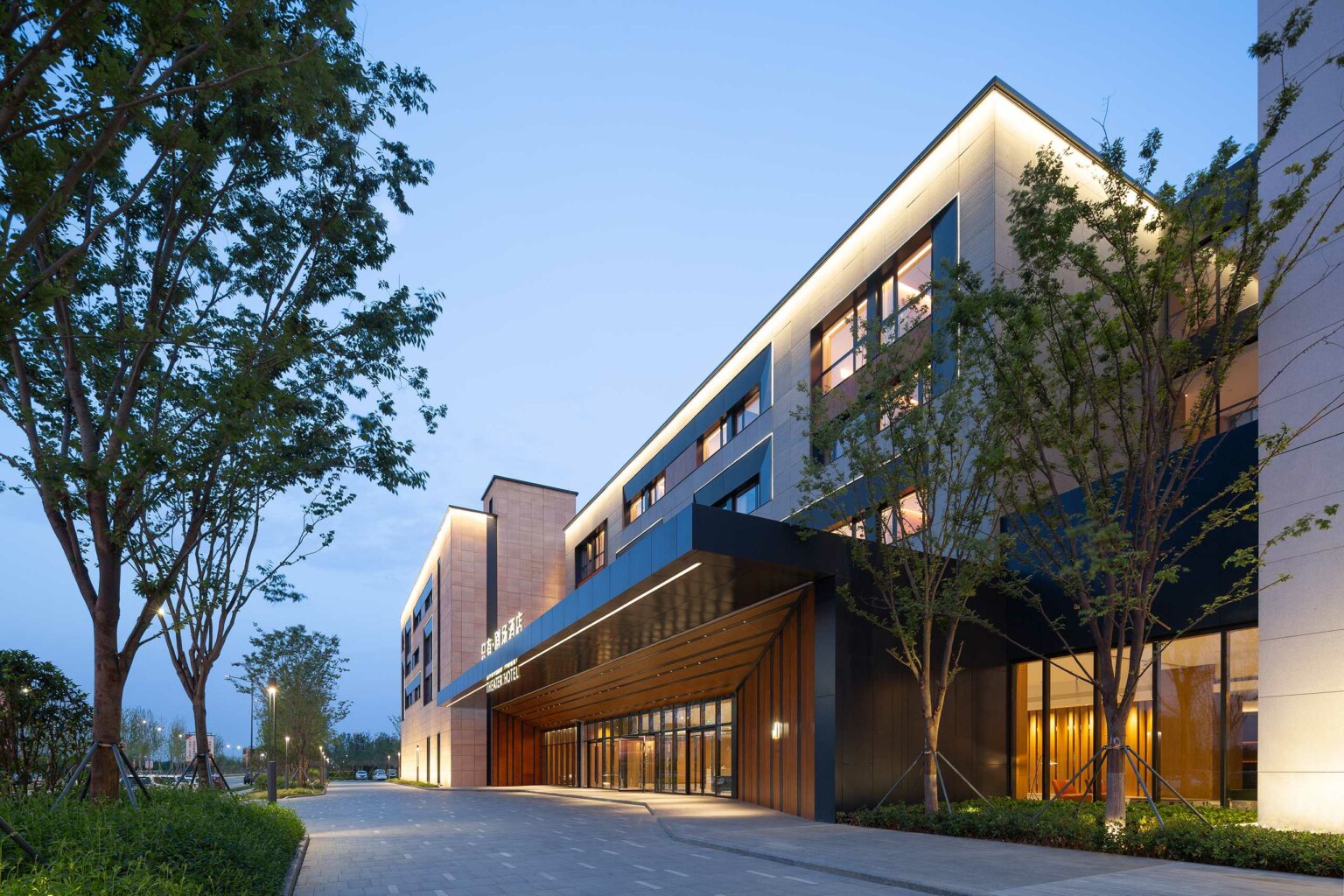 Modern hotel building at twilight with lit windows and a glass entrance. The sky is clear, and trees line the