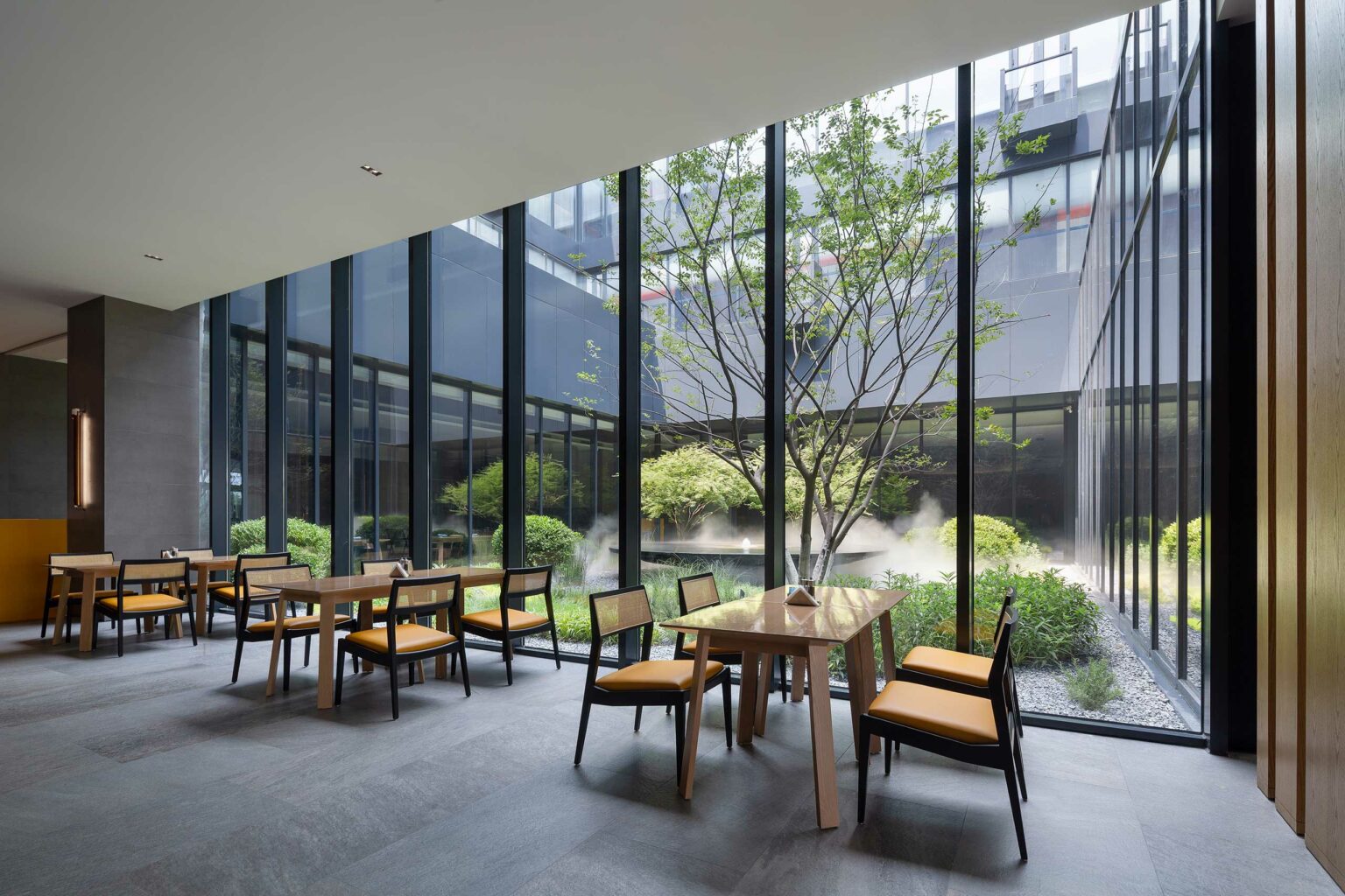 Modern hotel dining area with wooden tables and orange-cushioned chairs, adjacent to floor-to-ceiling windows overlooking a lush courtyard with trees.
