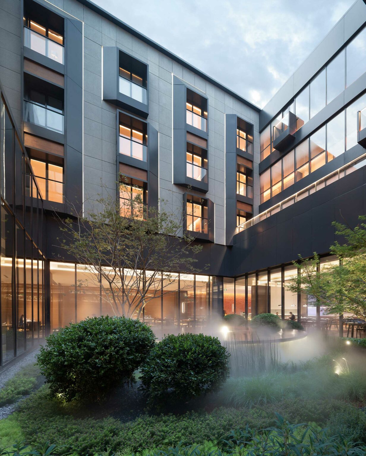 Modern hotel courtyard with sleek gray facade and large windows, warm interior lights glowing. Greenery and mist create a tranquil atmosphere.