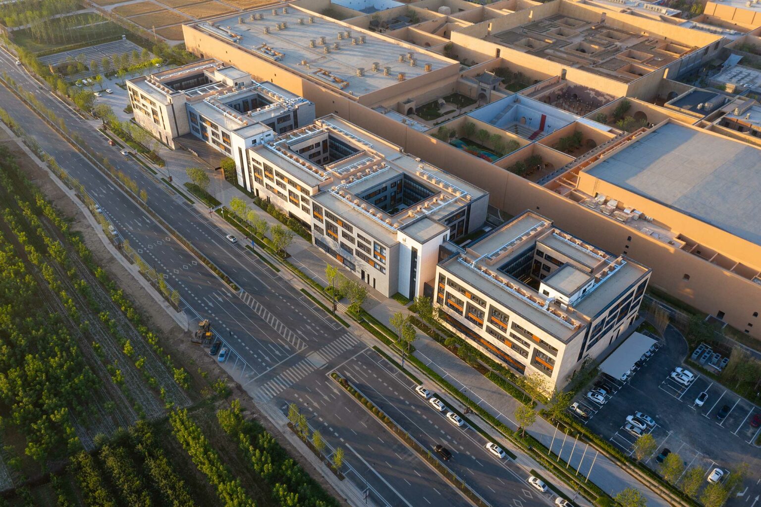 Aerial view of a modern industrial complex with a geometric design, adjacent to a wide, tree-lined street. The sun casts a warm glow, creating an organized, serene atmosphere.
