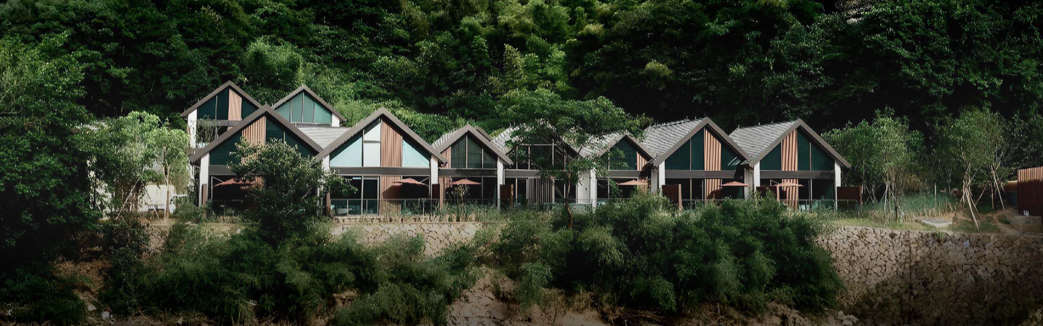 Modern cabins with A-frame roofs are nestled against a lush green hillside, overlooking a gentle river. The scene conveys tranquility and natural beauty.