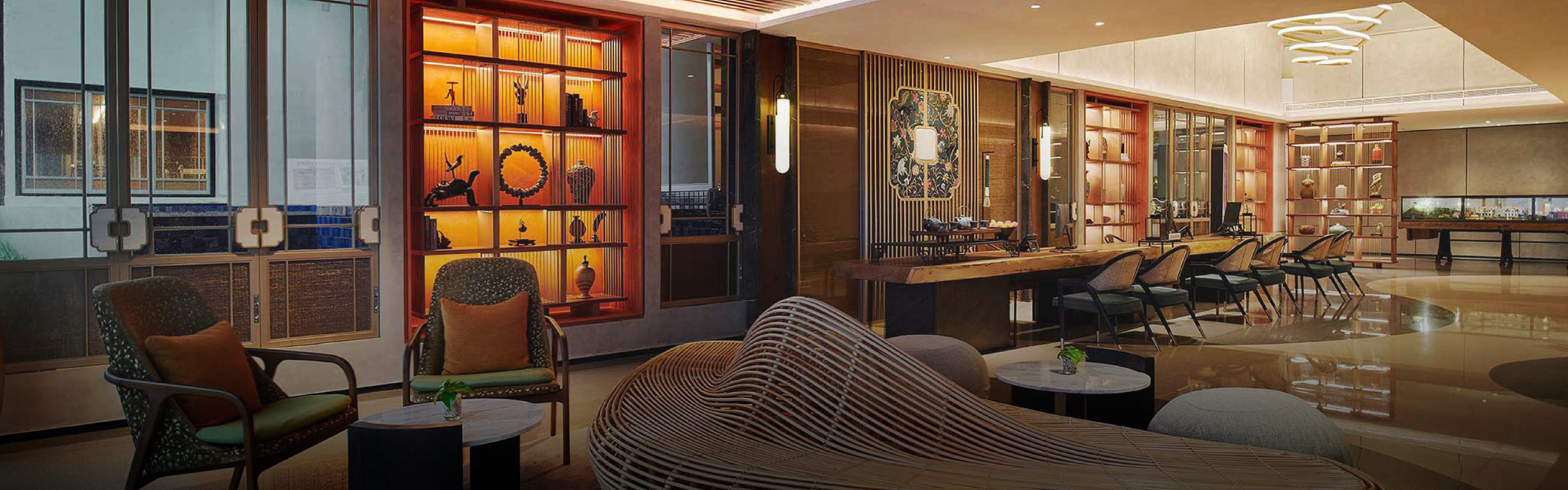 Modern hotel lounge with curved wooden seating, elegant armchairs, and a backlit shelf displaying art. Warm lighting creates a cozy, sophisticated ambiance.