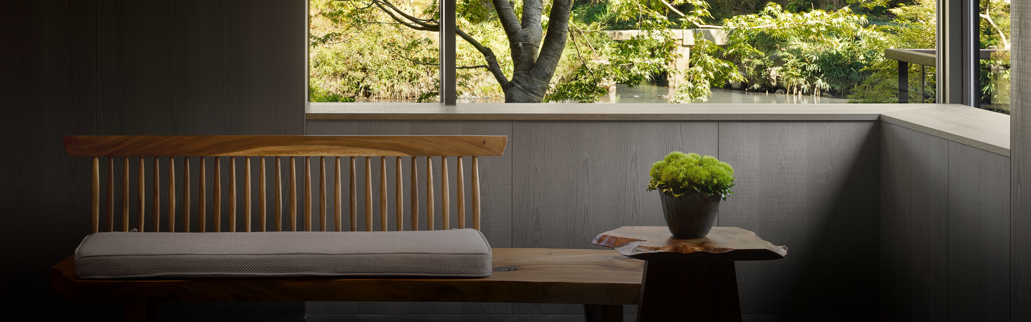 Calm interior hotel space with Japanese garden view. Wooden furniture, patterned rug, and bonsai in foreground; lush greenery and rocks seen through large windows.